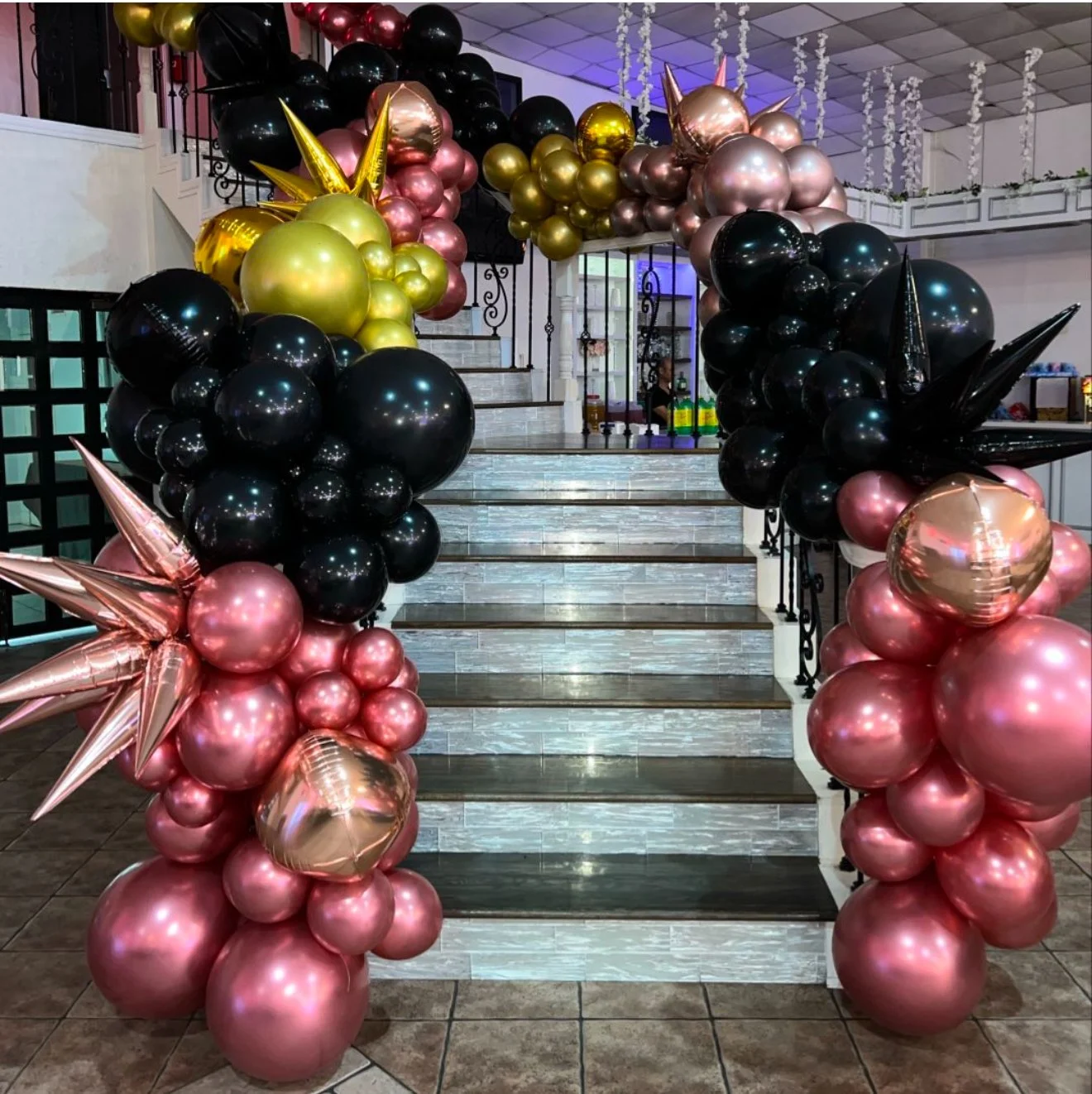 Decorative balloon arch with pink, black, gold, and rose gold balloons at the base, and more pink and gold balloons on top, arranged at the entrance of a staircase.