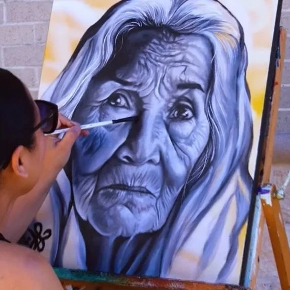 Artist painting a detailed portrait of an elderly woman with long, white hair on a large canvas.