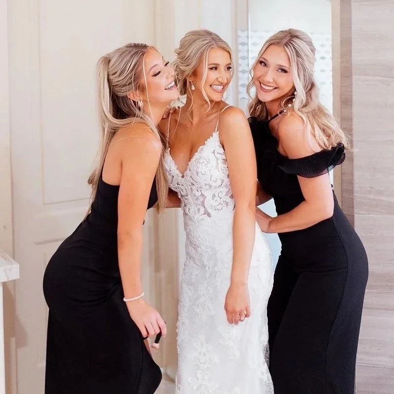A bride in an elegant white lace wedding dress with off-the-shoulder long sleeves, standing outdoors with a garden and orange wall in the background.