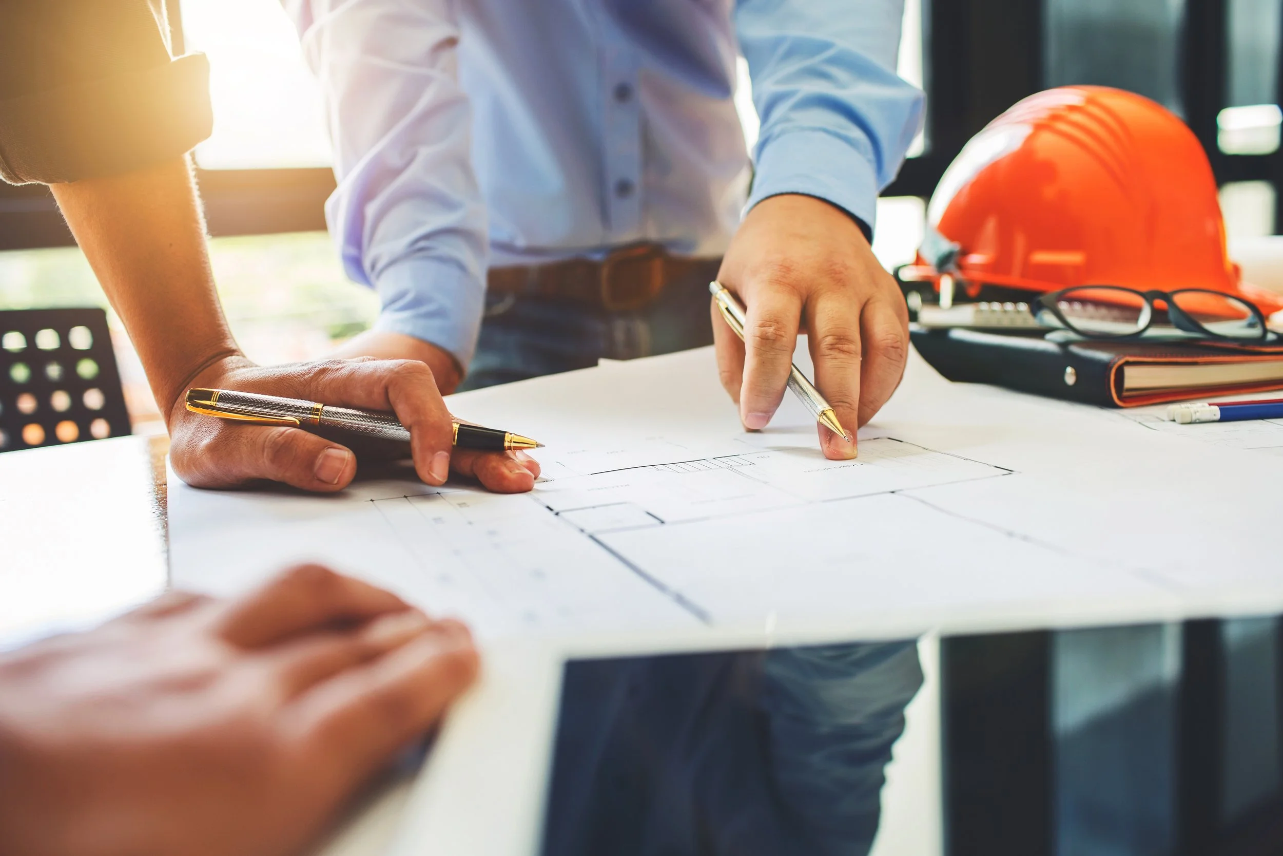 Person with a yellow and black pen pointing at blueprint on desk, orange safety helmet, glasses, and notebook nearby.