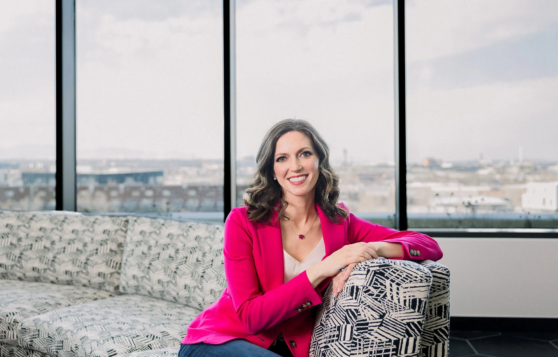 A woman with long, wavy brown hair wearing a bright pink blazer and white top sits on a patterned armchair in a modern, high-rise office with large windows showing a cityscape and cloudy sky.