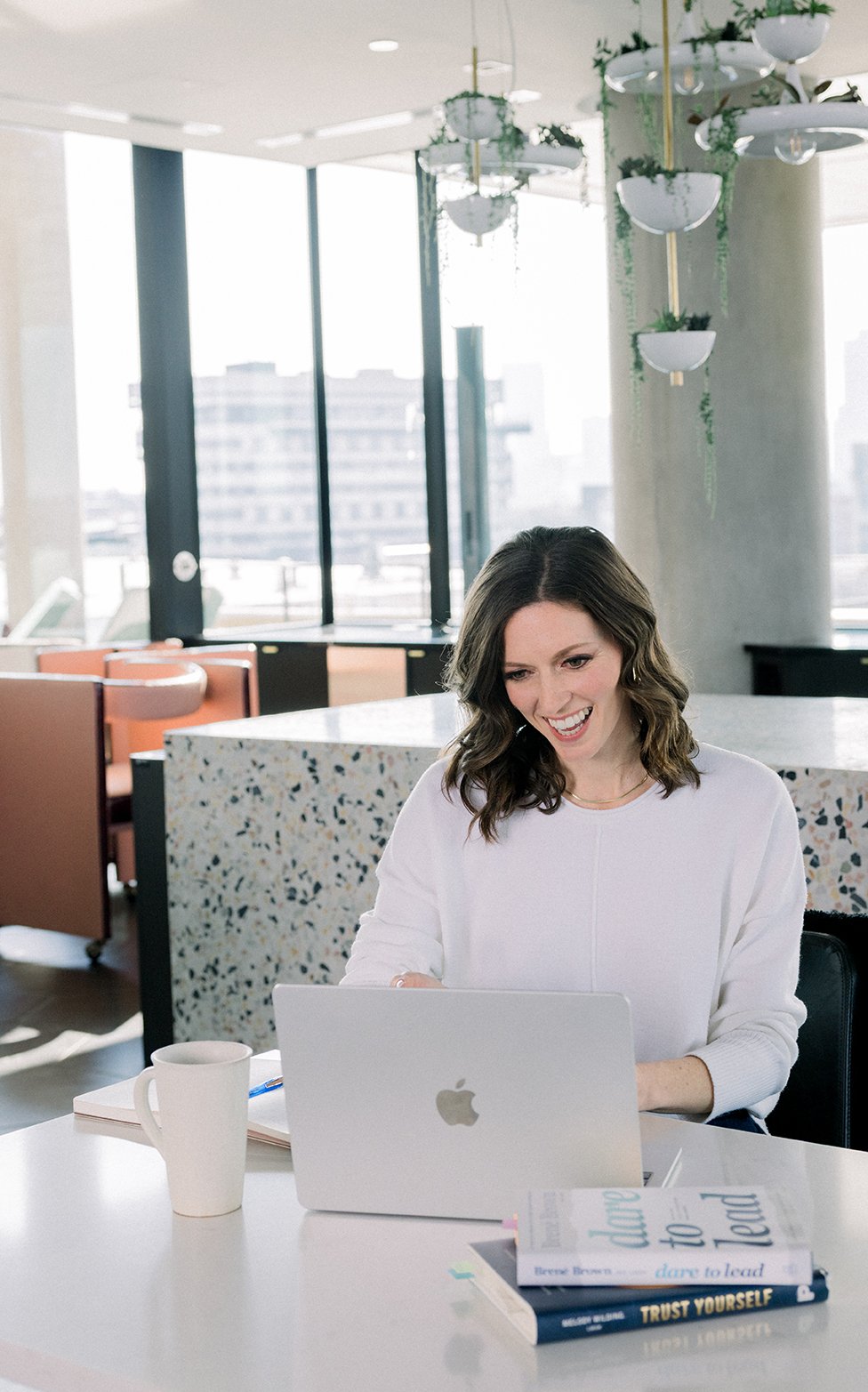 A woman with shoulder-length wavy brown hair smiling at her open silver MacBook on a white table in a bright, modern office space with large windows, books, a mug, and a notebook.