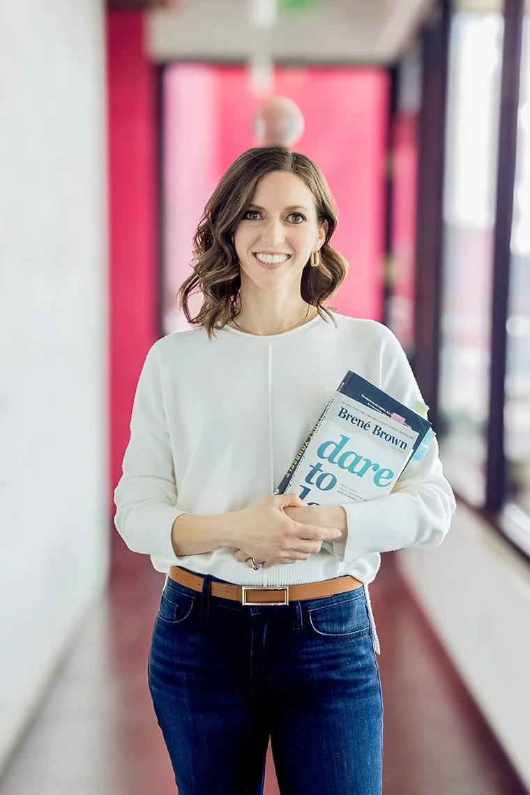 A woman with shoulder-length brown hair wearing a white sweater and jeans, holding books, standing in a brightly lit hallway with pink and black walls and large windows.