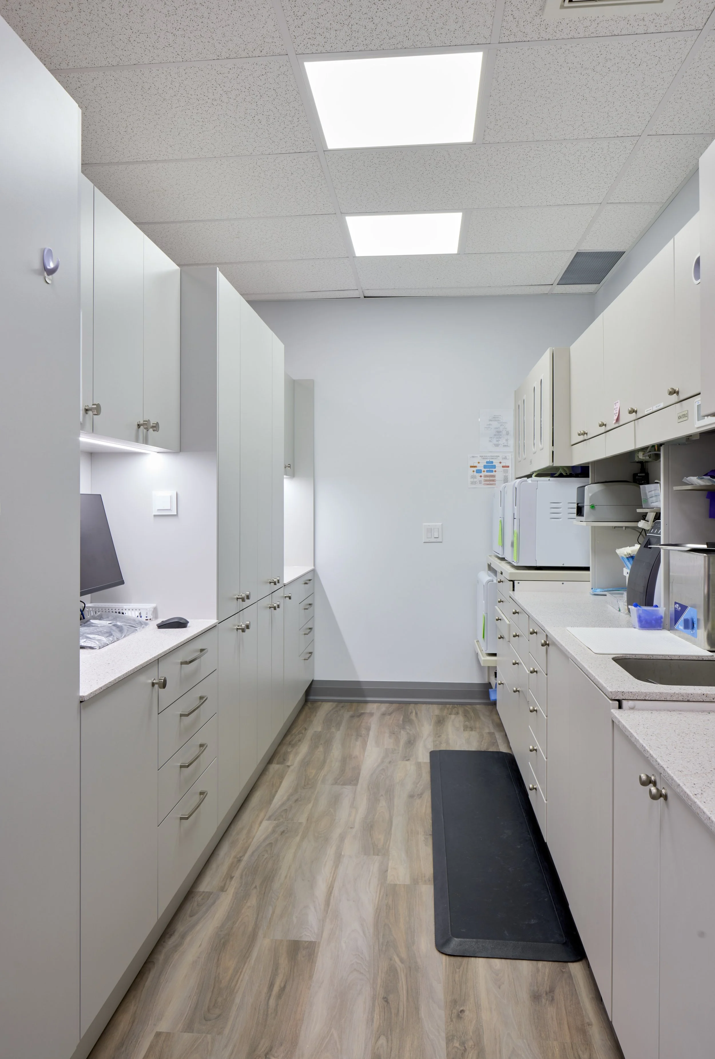 Dental or medical office kitchen with white cabinets, countertops, and a black floor mat on light wood flooring, under a ceiling with fluorescent lights.