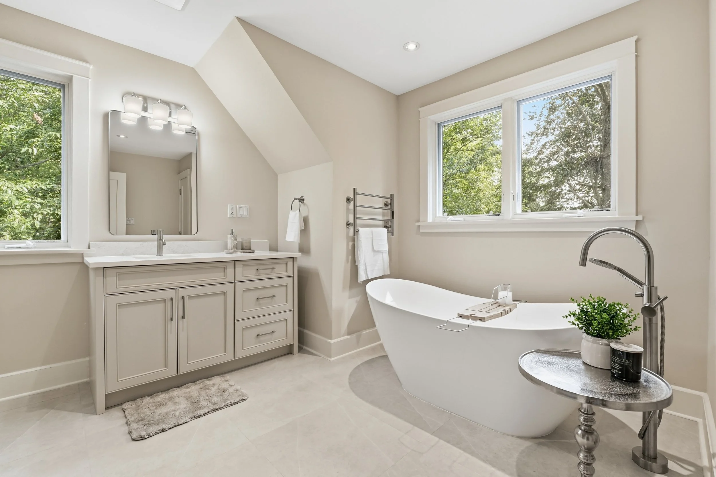 Bright bathroom with large windows showing green trees outside. Features include a white vanity with a mirror, bathtub, and towel rack. Decor includes a small potted plant and a rug.