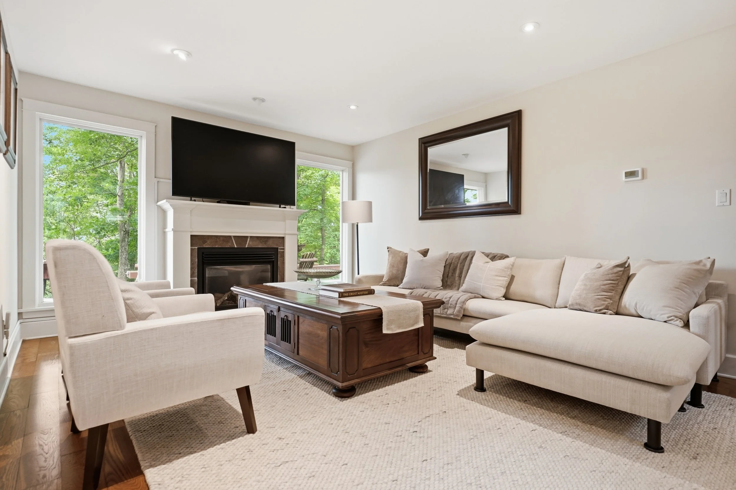 Living room with large beige sofa, white armchair, wooden coffee table, fireplace, TV above fireplace, large mirror on wall, windows showing green trees outside, floor lamp, carpeted floor.