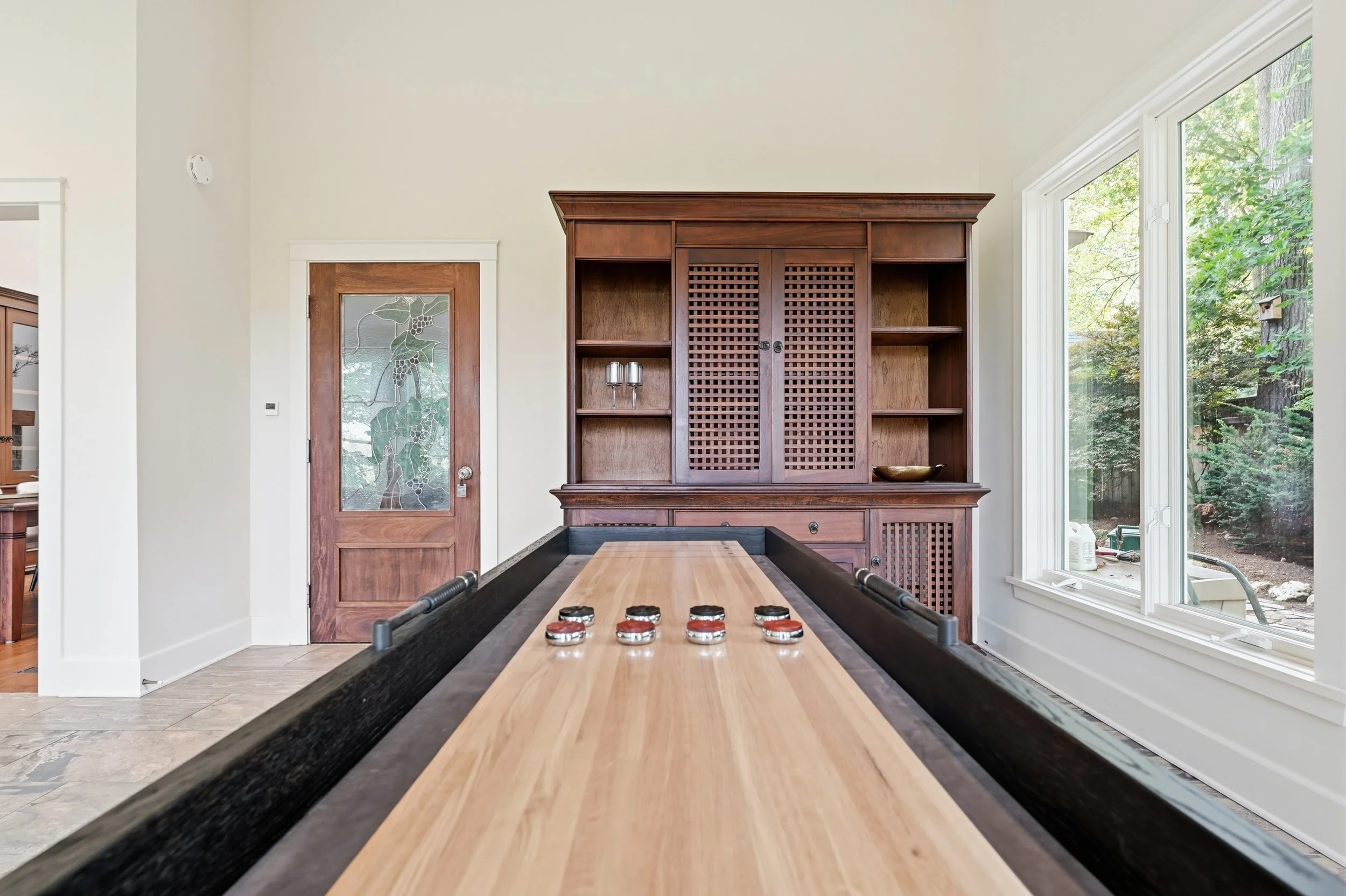 Interior view of a room with a wooden shuffleboard table, a large wooden cabinet with lattice doors, a door with decorative glass, and a large window showing trees outside.