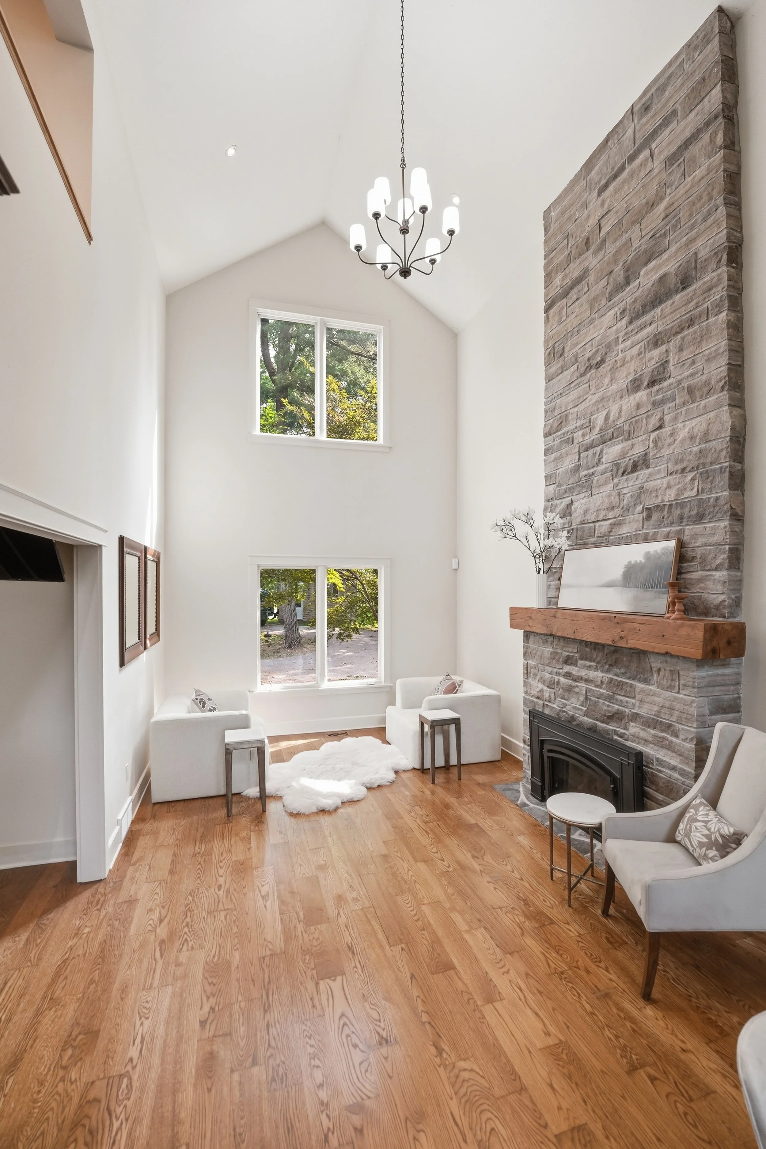 Living room with wooden flooring, white walls, large window, stone fireplace with wooden mantel, chandelier, and chairs.