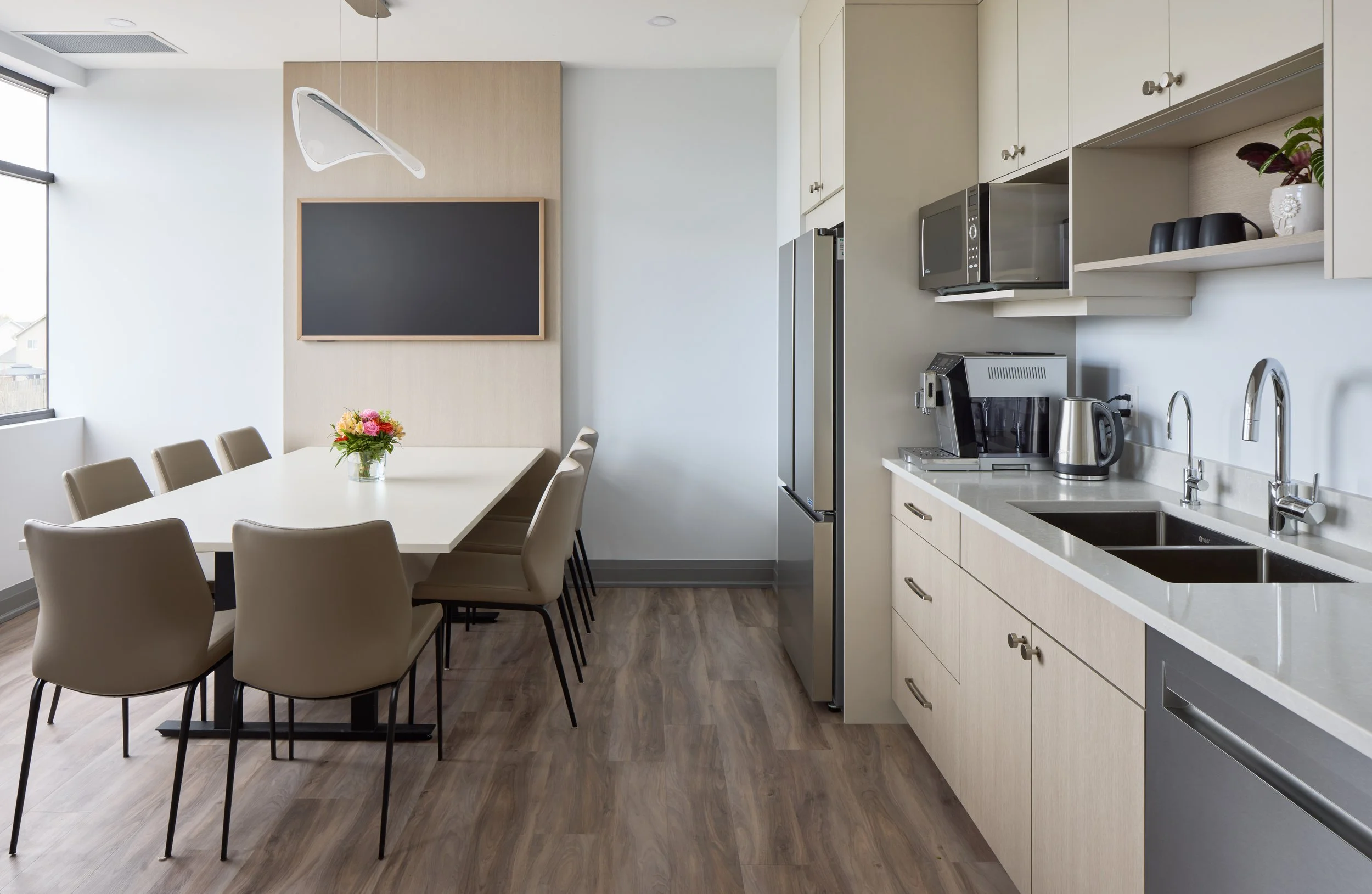 Modern kitchen and dining area with white walls, wooden flooring, a long white dining table with beige chairs, a vase of pink flowers, a blackboard on the wall, and kitchen appliances including a microwave, refrigerator, coffee machine, and kettle.
