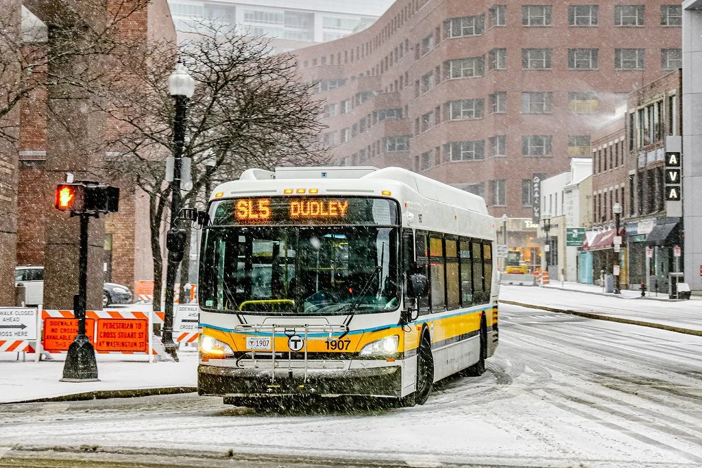 A photo of a city bus, with "SL5: Dudley" on the marquee, turning a corner in snowy Boston