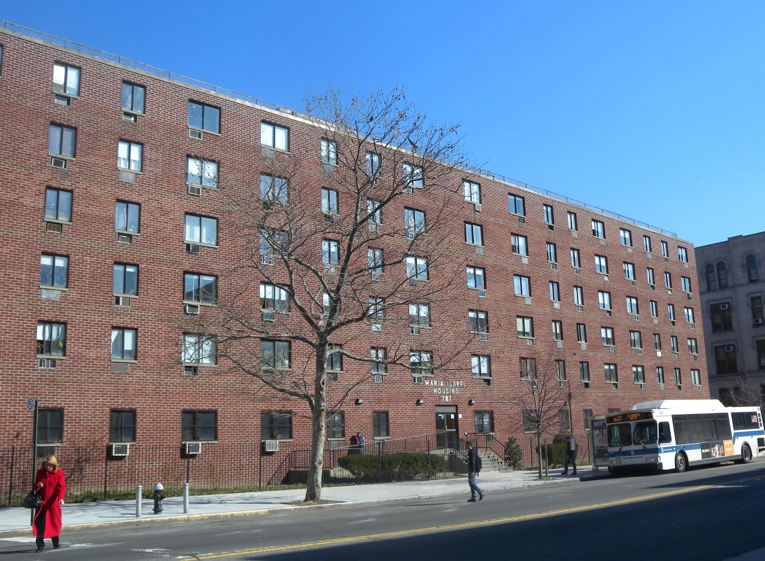 A photo of the facade of a large, brick residential building under a blue sky, with people walking in front of it.
