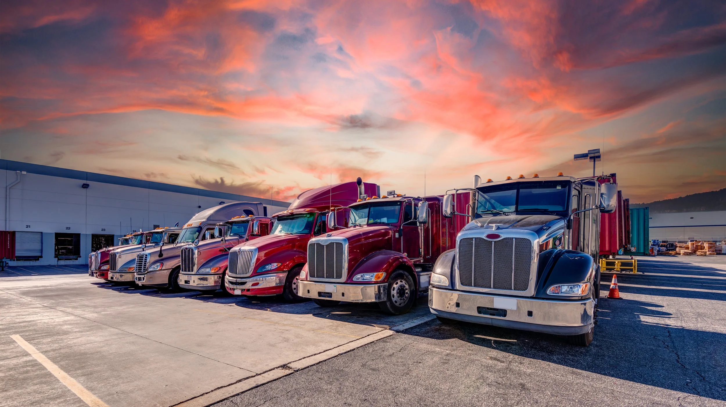 Row of parked semi-trucks at a warehouse under a vibrant sunset sky.