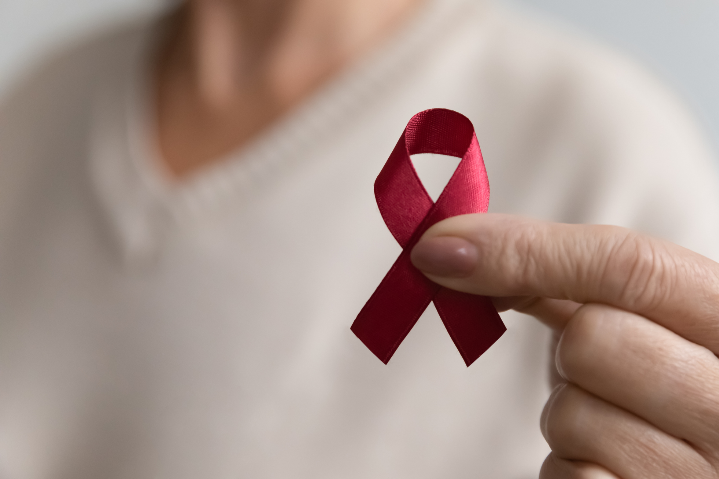 Person holding a red awareness ribbon between their thumb and index finger.