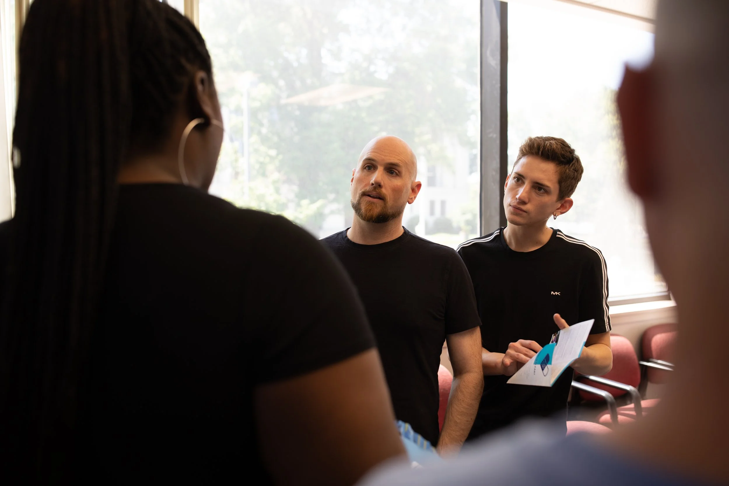 A diverse group of people engaged in a discussion during a meeting or workshop, with natural light coming through large windows.