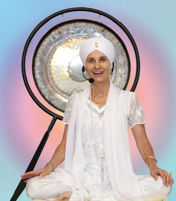 A smiling female yoga teacher wearing a white turban and traditional white clothing, sitting cross-legged in front of a gong. She is wearing a headset and appears to be speaking.