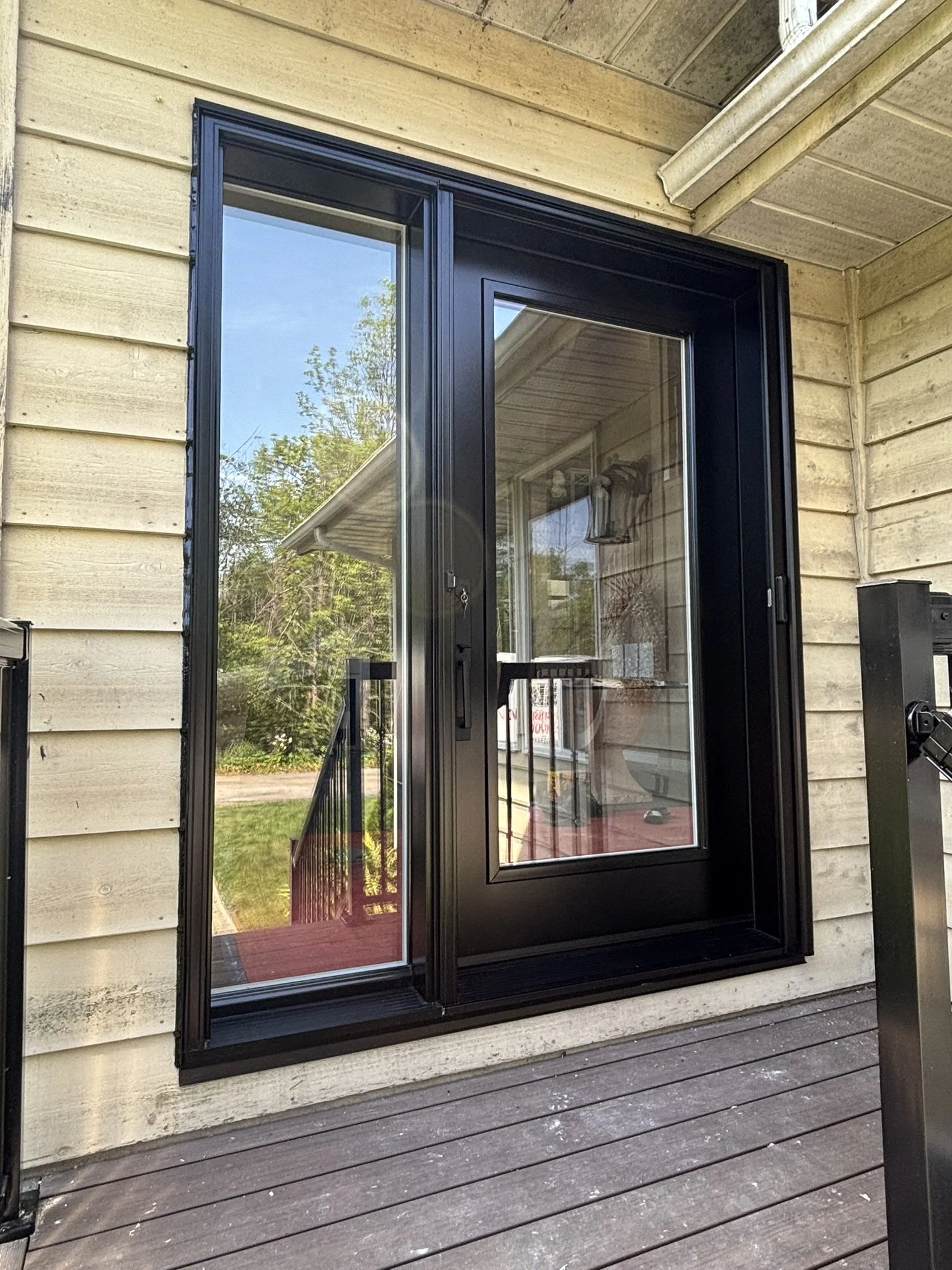 Black framed sliding glass door on a wooden deck outside a house with beige siding and a porch light inside.