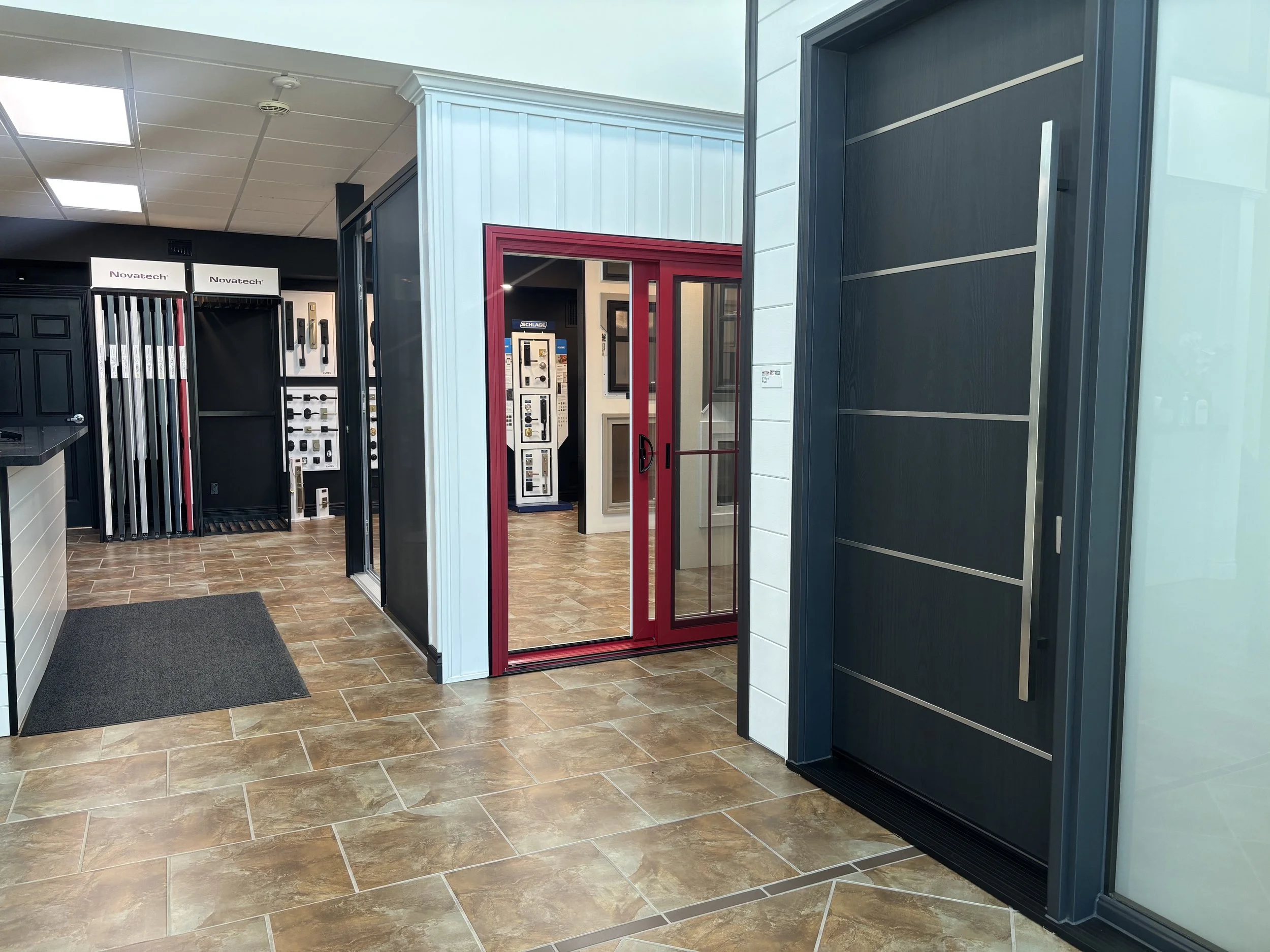 Showroom with display of window and door samples, featuring a red door frame, a black display with hardware, and tile flooring.