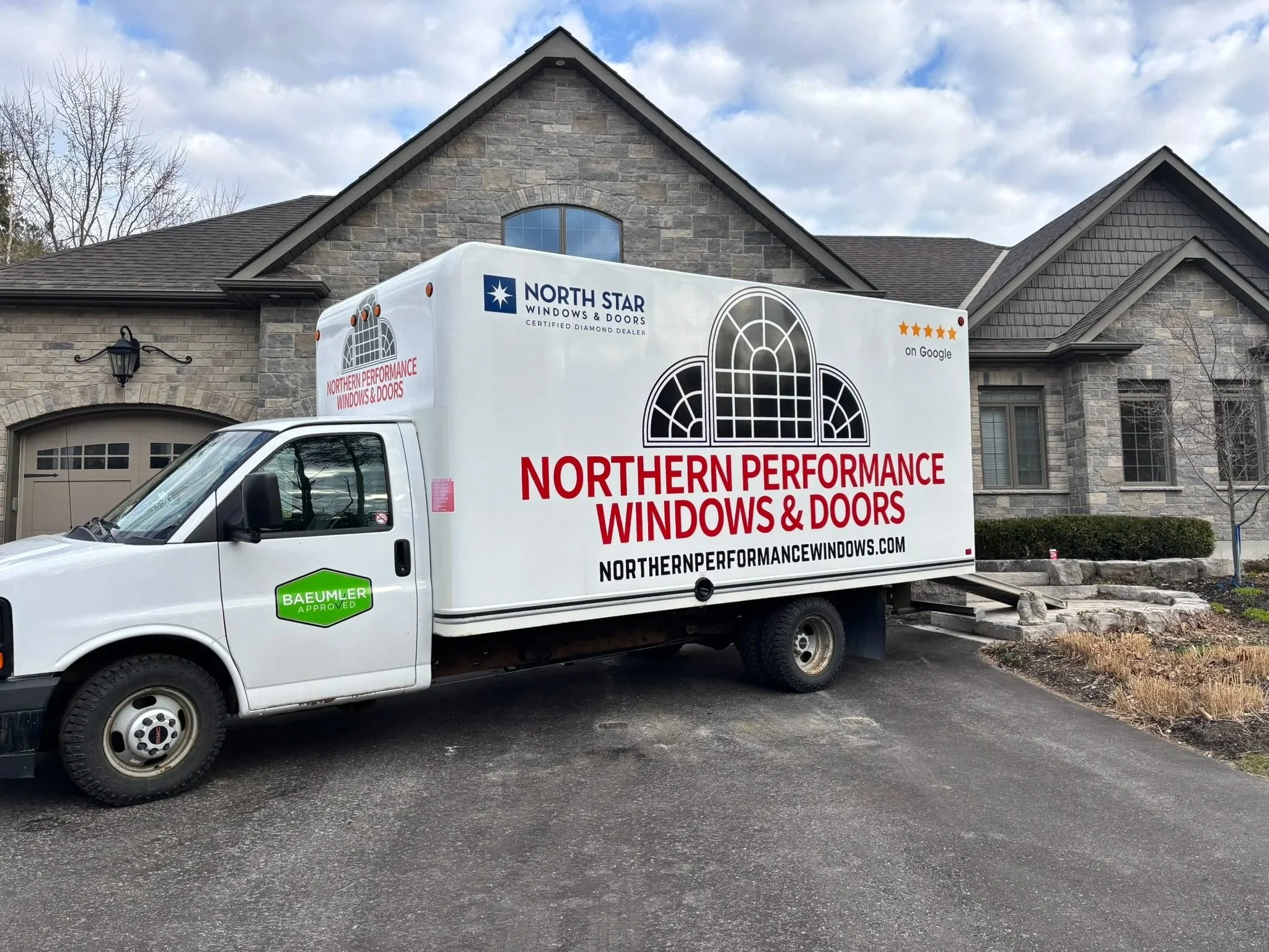 A white work truck parked in front of a brick house under a cloudy sky. The truck has signage for Northern Performance Windows & Doors, featuring a window graphic and logos, including North Star Windows & Doors and Baumlser Approved.