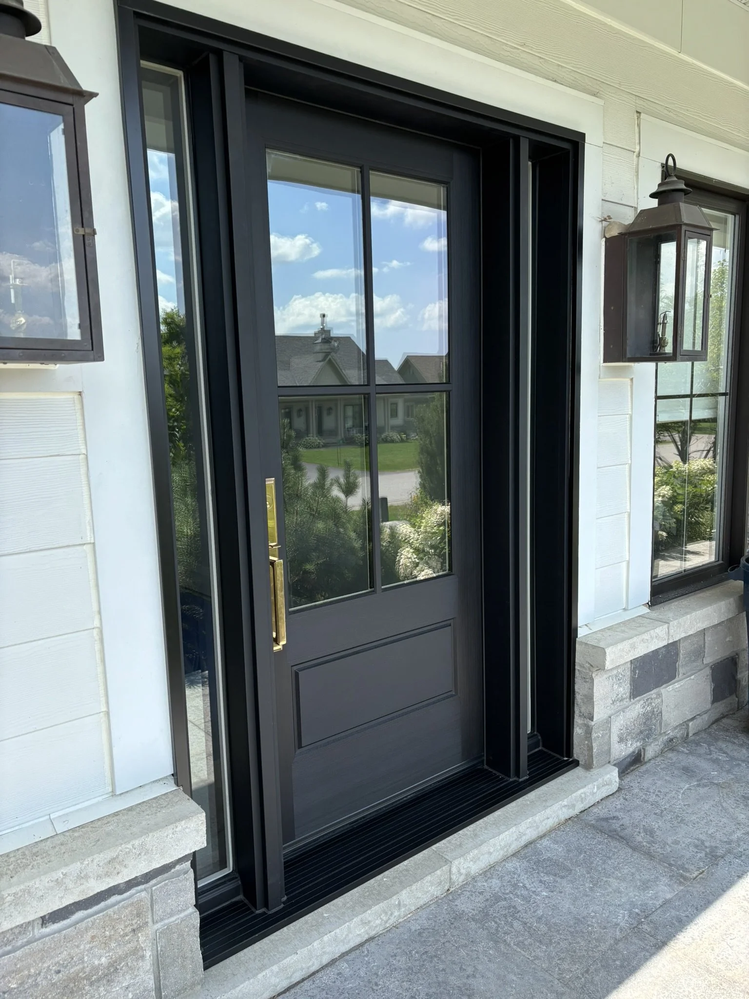 Front door with glass panels, dark gray color, and brass handle, flanked by two wall-mounted lantern-style outdoor lights on a porch with stone flooring and white siding, with a view of a neighborhood street and houses in the reflection.