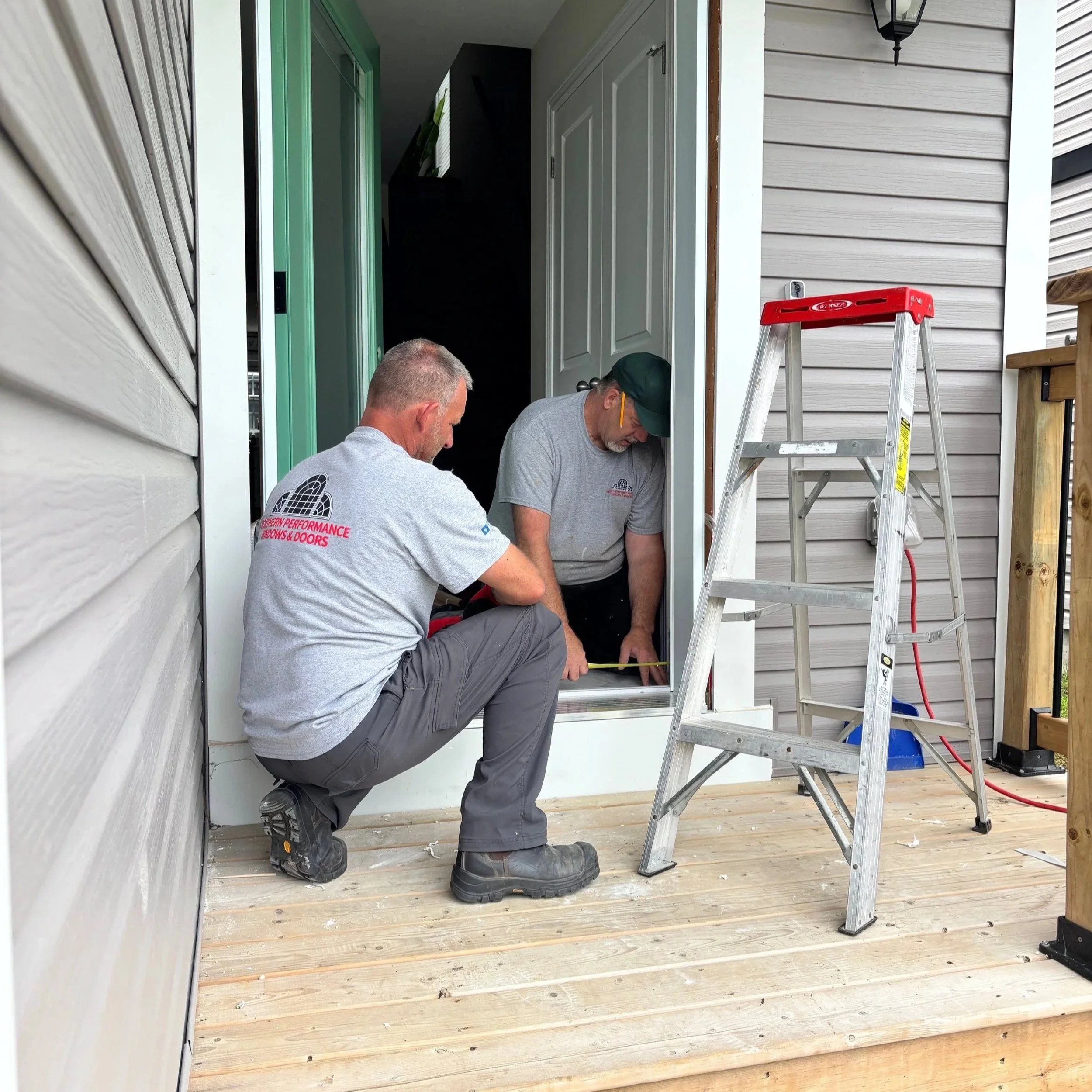 Two men working on the installation or repair of a door frame on a house porch. One man is crouching and looking down, the other leaning forward, both focused on the task. There is a ladder and a worksite with tools nearby.