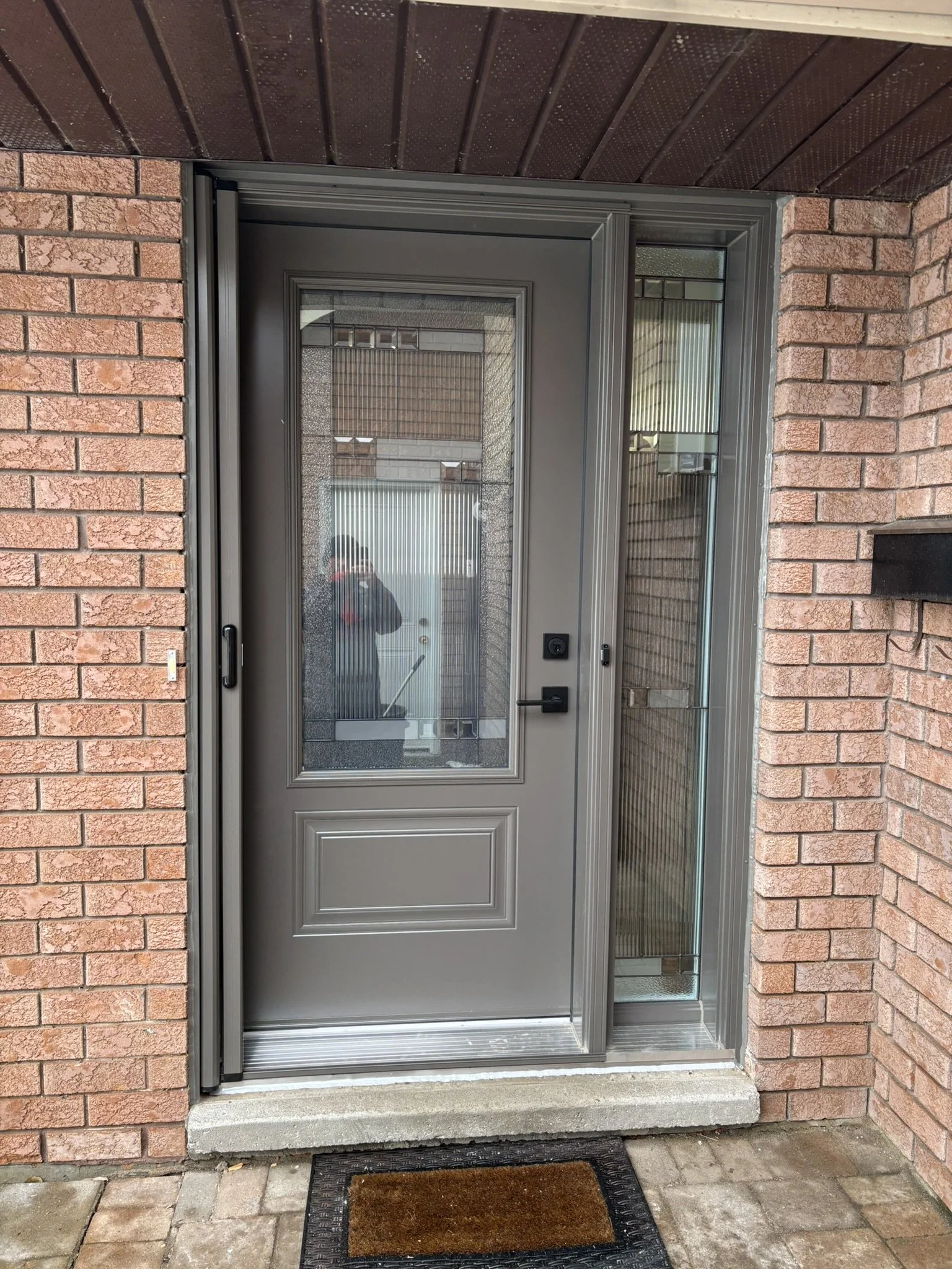 A front door with a glass panel, extra glass side panels, and a black doorbell, set in a brick wall with a brown doormat in front.