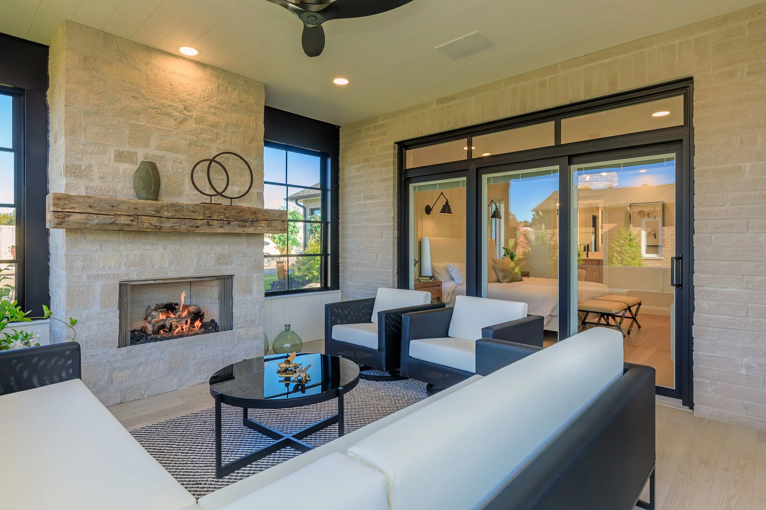 Living room with a stone fireplace, modern furniture, large windows showing outdoor greenery, and a bedroom visible through glass doors.