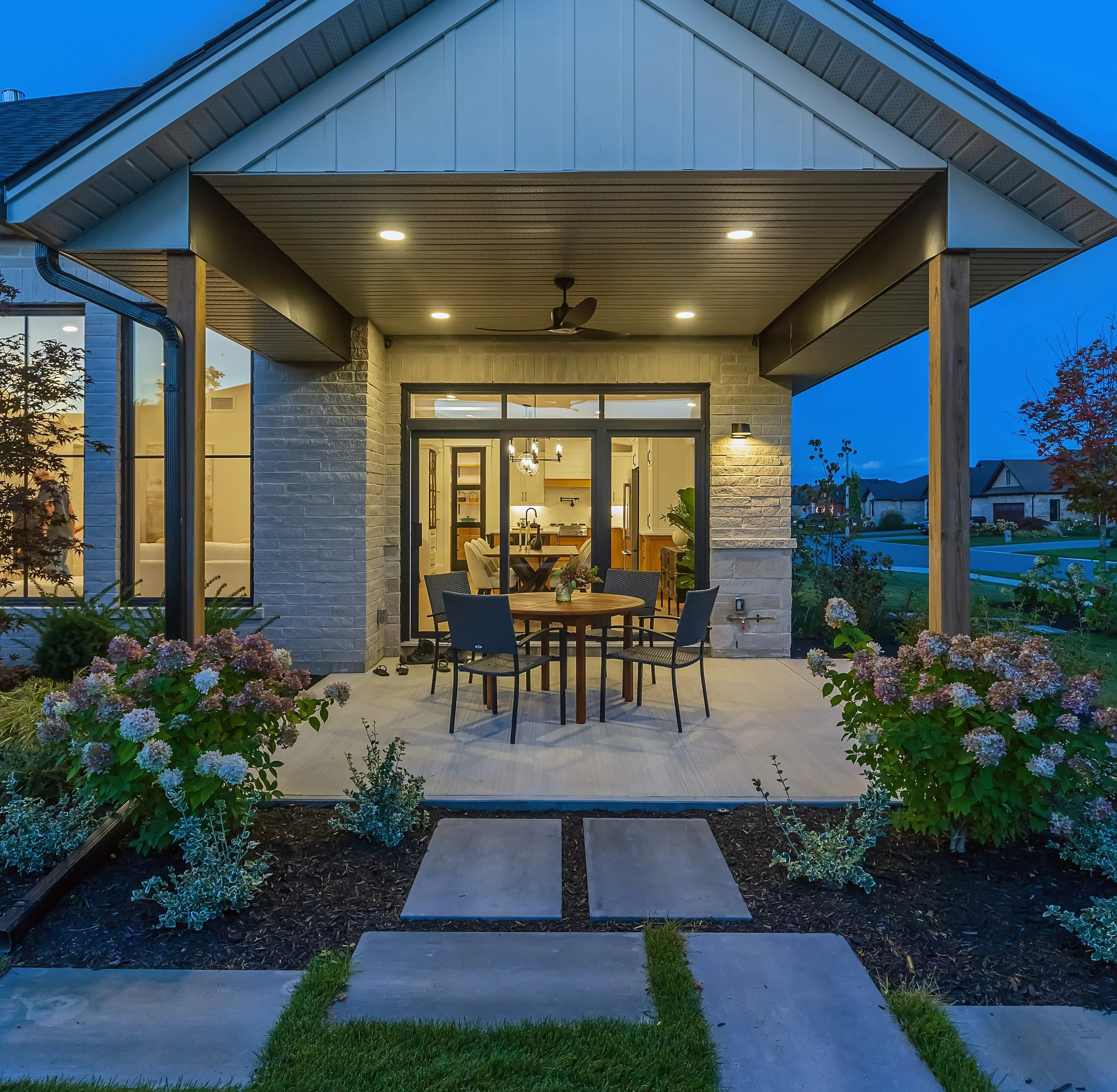 The image shows the back porch of a house at dusk. The porch is lit with four ceiling lights and has a ceiling fan. There is a dining table with chairs in the background, and the house features large glass doors and windows. The porch is surrounded by a garden with flowering bushes and a concrete walkway.
