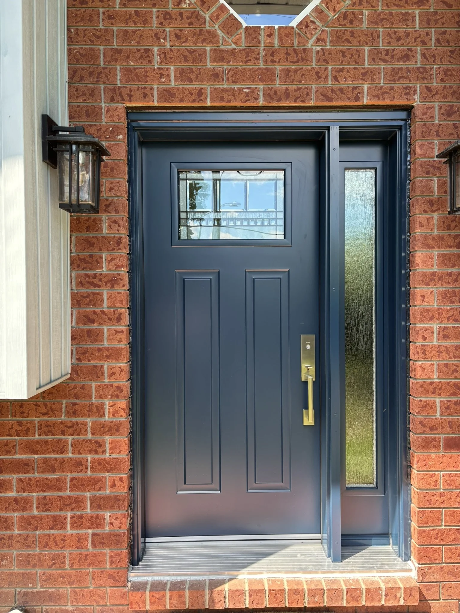 Close-up of a modern dark blue front door with a small rectangular window, surrounded by red brick walls and two outdoor wall-mounted lanterns on either side.
