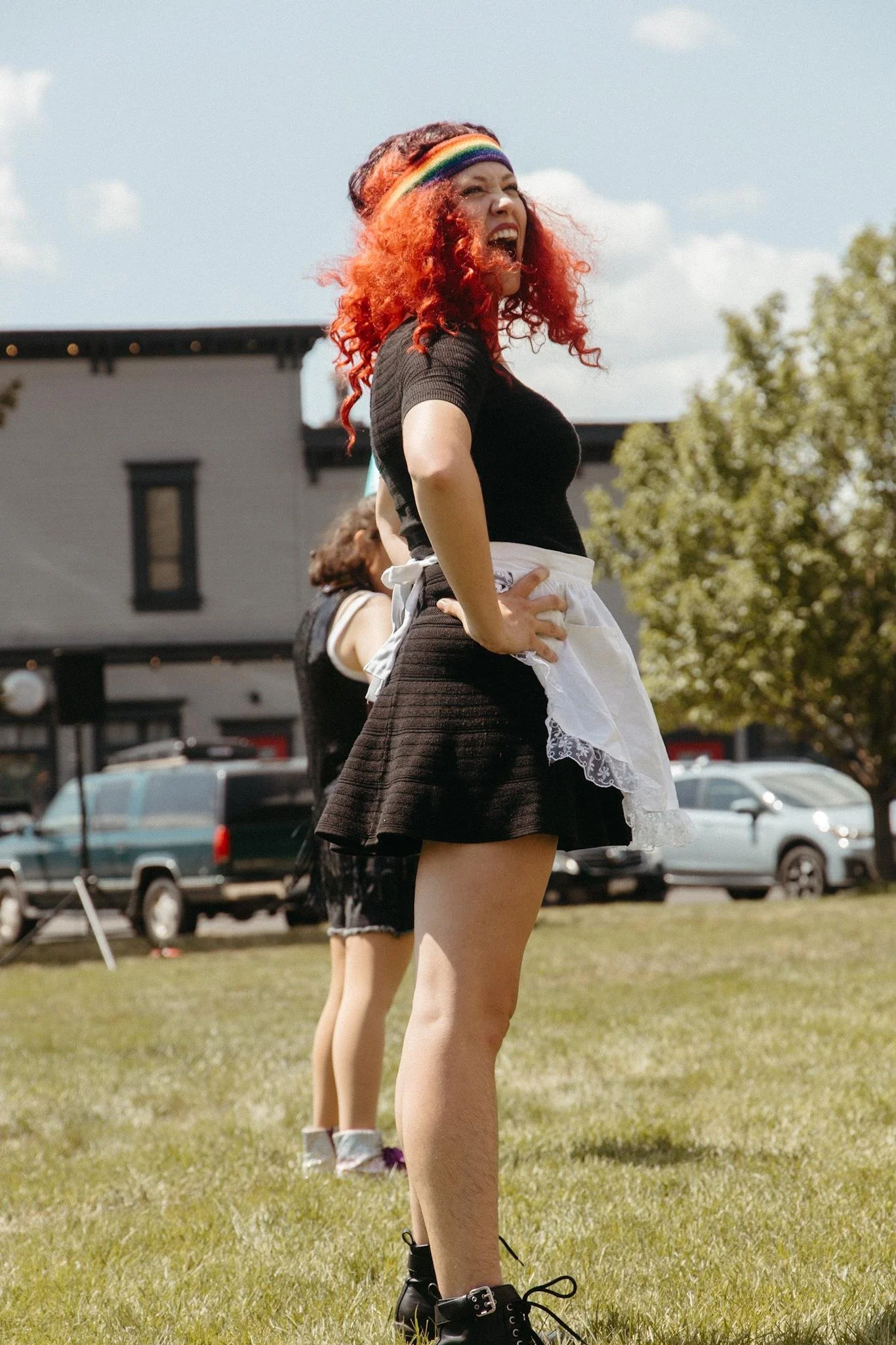 A woman with curly red hair, wearing a rainbow headband, black top, and black skirt, laughing outdoors on a sunny day, with her hands on her hips, and another person partly visible behind her.