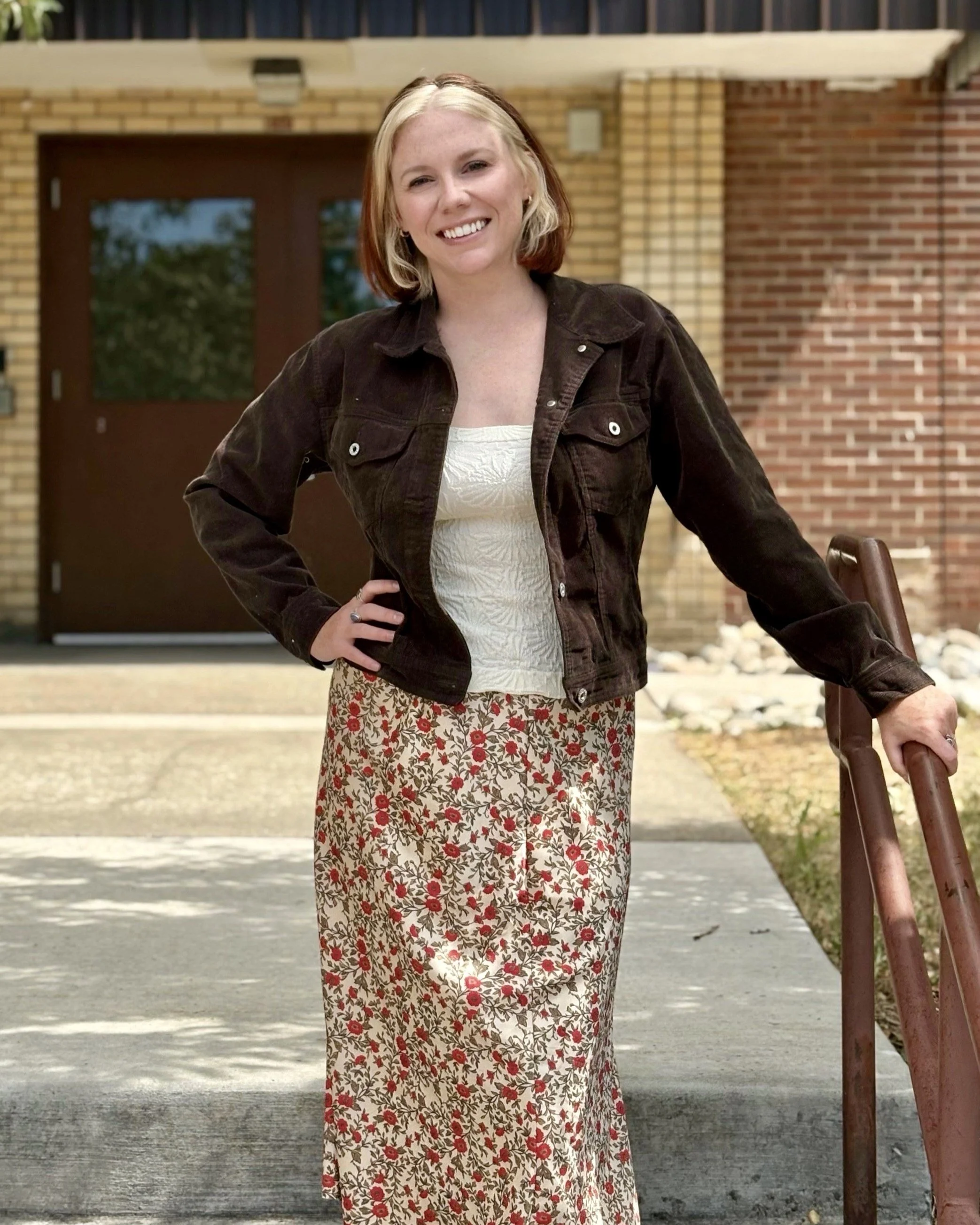 A woman with shoulder-length blonde hair, wearing a dark brown jacket over a white top, standing outside on a concrete path with a brick building in the background, smiling and holding a railing.