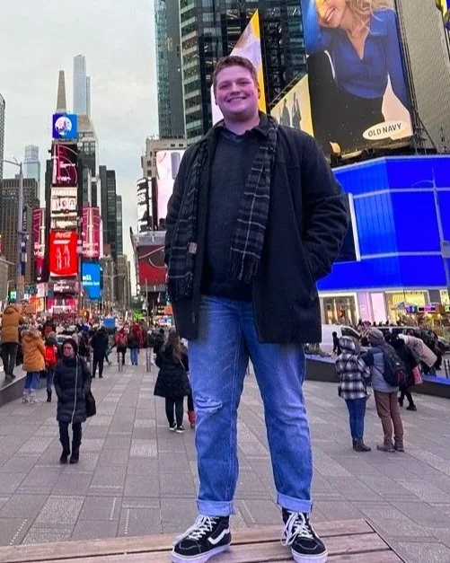 A young man smiling in Times Square, New York City, surrounded by illuminated billboards and people walking.