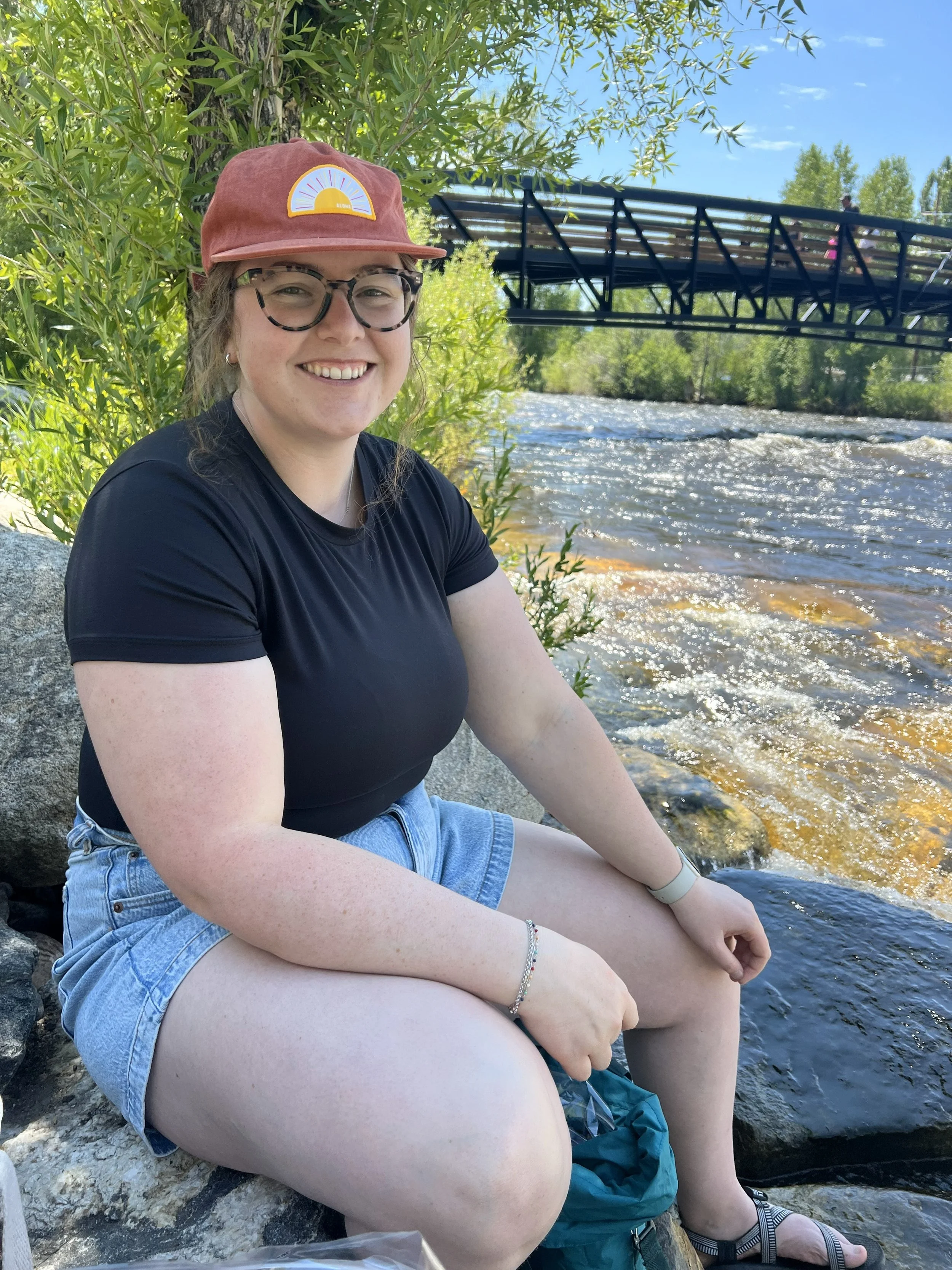 A young woman sitting on rocks by a river, smiling at the camera, with a bridge in the background and surrounded by green foliage.