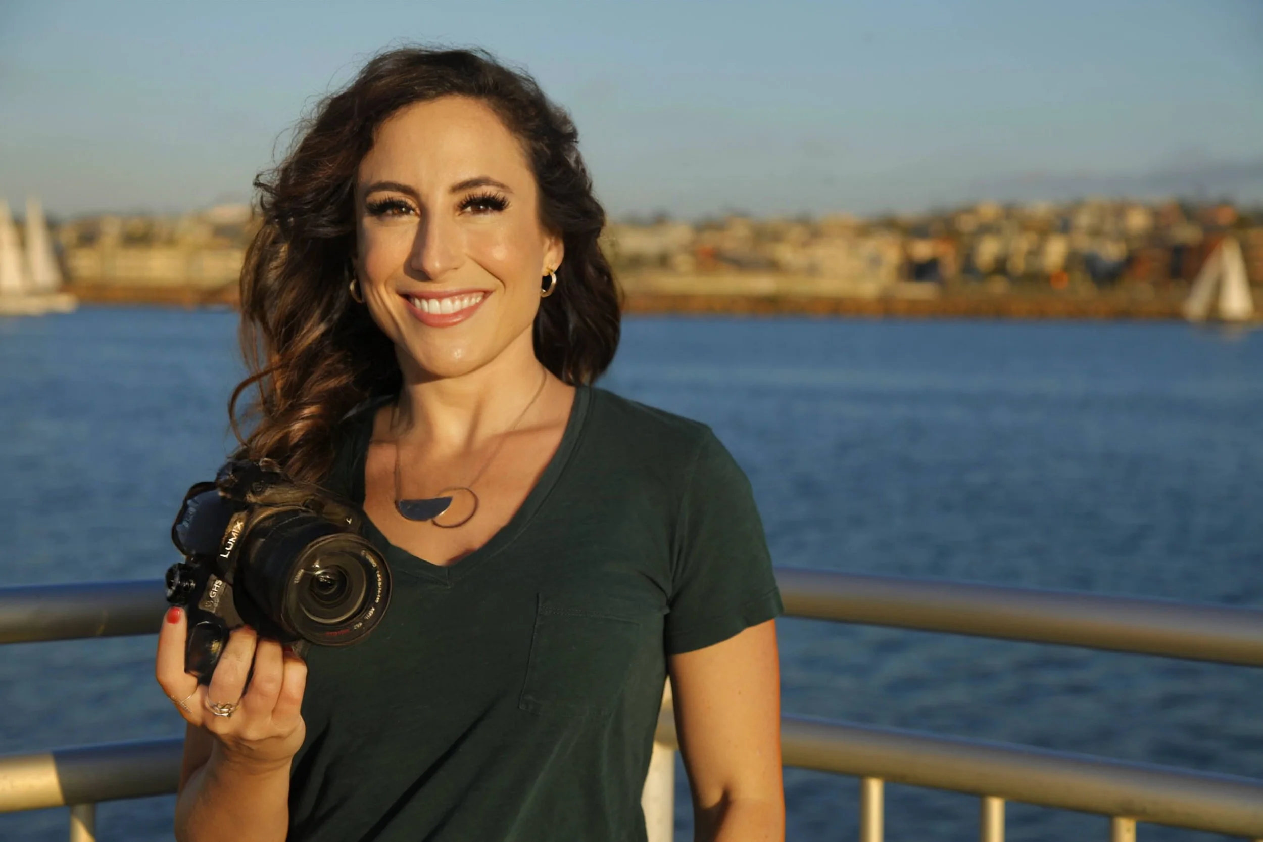 A smiling woman with wavy brown hair holding a camera outdoors by a body of water, with sailboats and buildings in the background during sunset.
