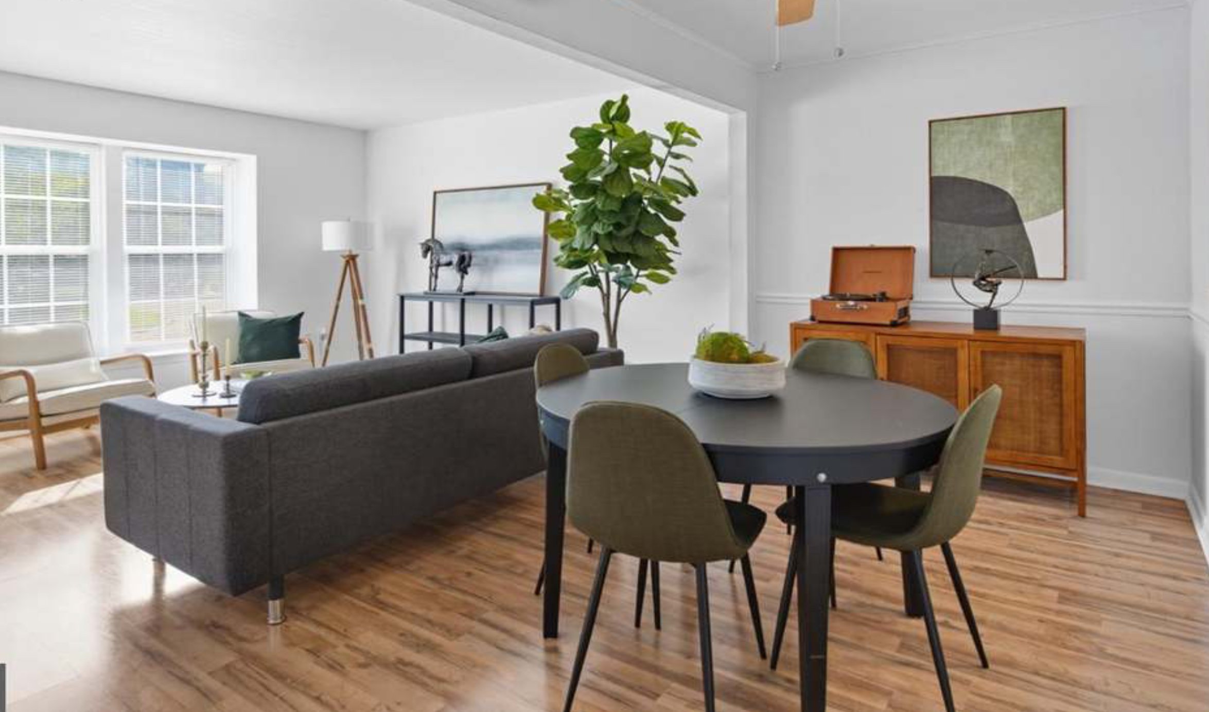 A living room with a round dining table and six chairs, a gray sofa, a white armchair, a large houseplant, a sideboard with a vintage record player, and framed artwork on white walls with large windows.