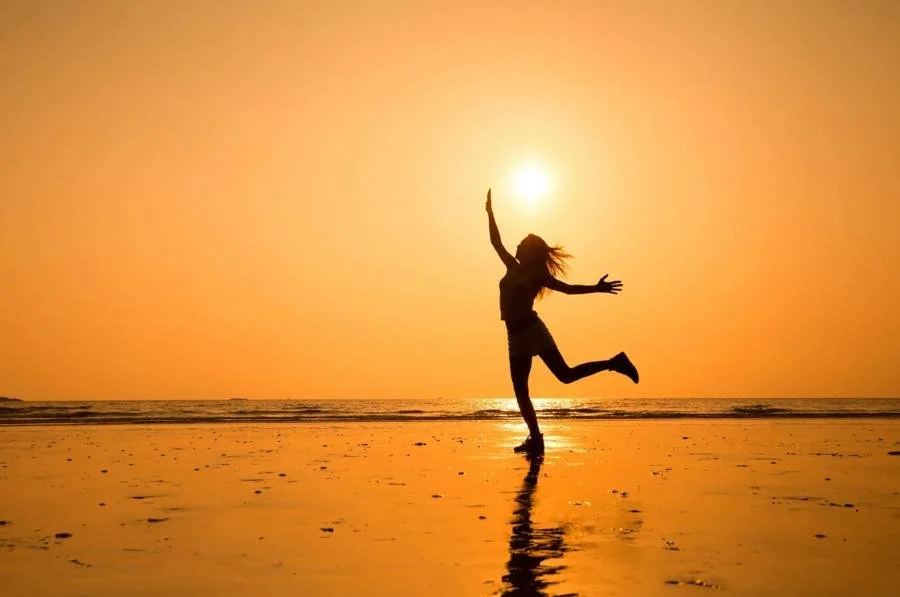 A joyful person dancing on the beach at sunset, arms raised toward the sun