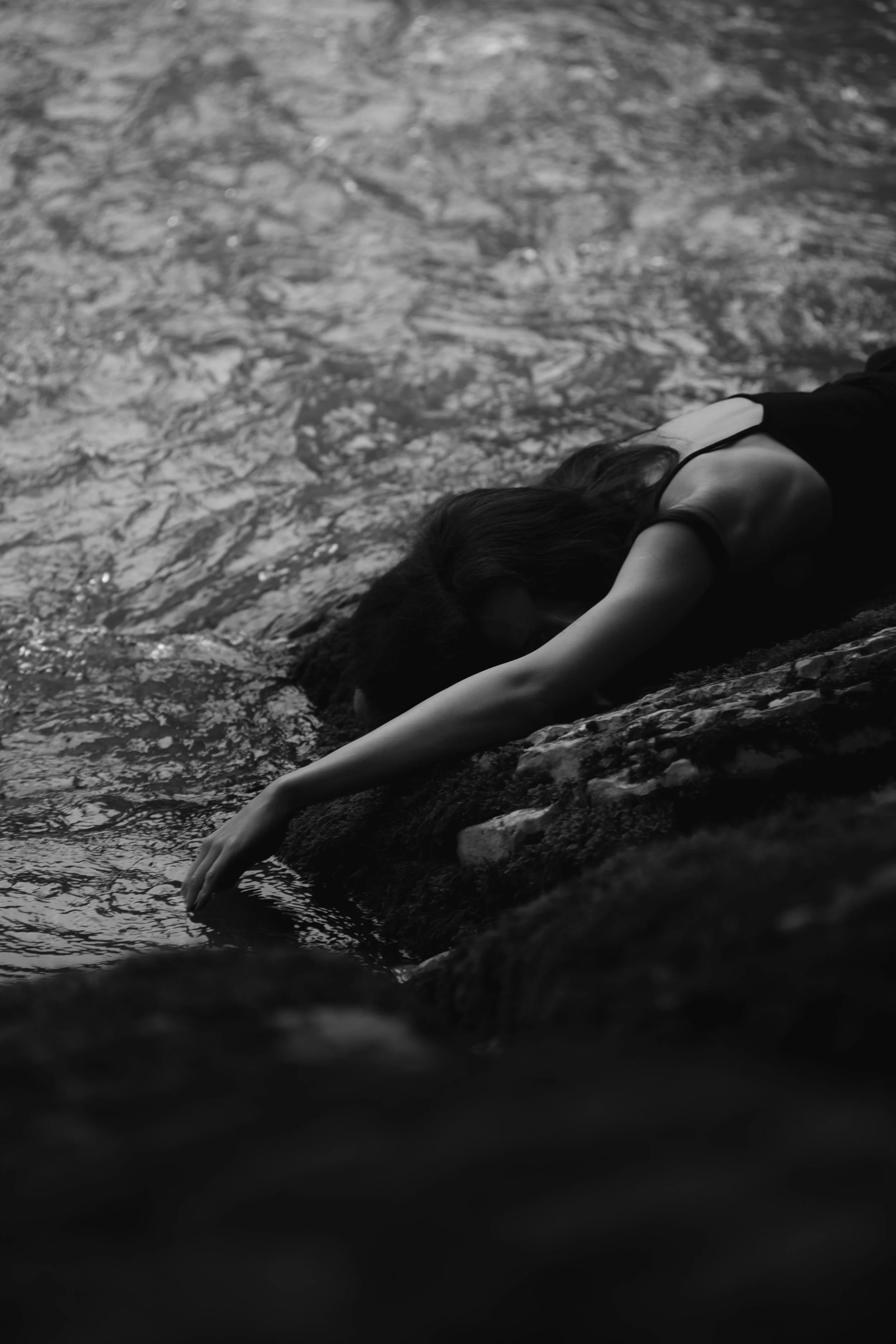A woman with wavy brown hair wearing a white sleeveless top, standing outdoors among trees.