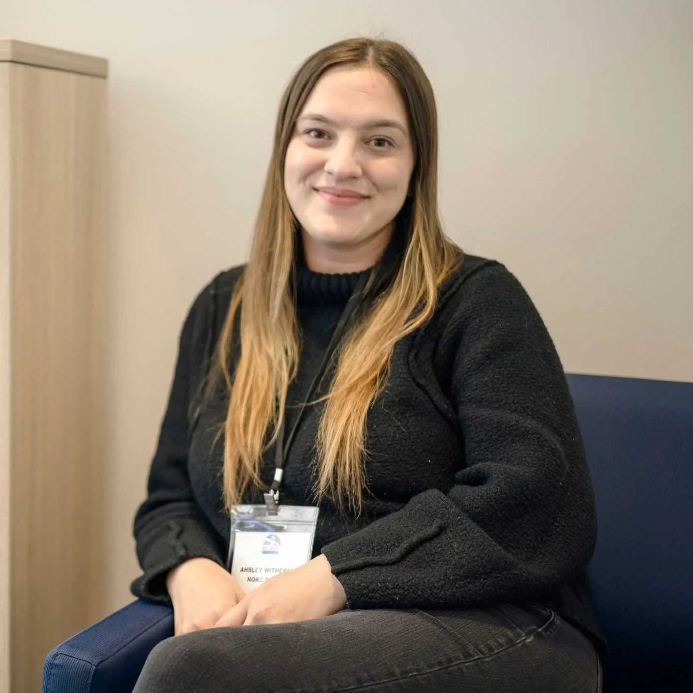 A person with long hair sitting on a chair, wearing a black sweater and a visible name badge.