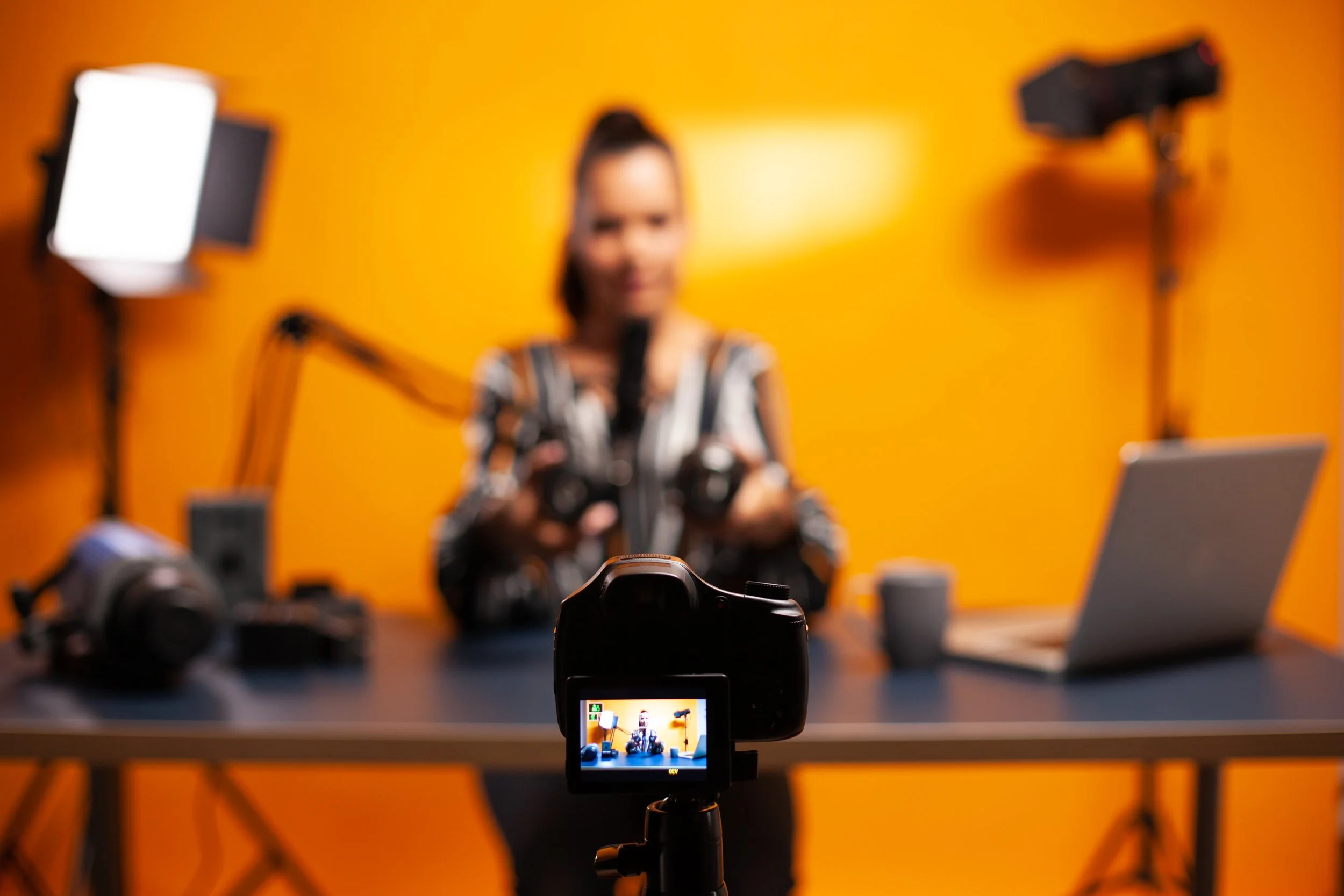 A woman sitting at a desk with video recording equipment, including a camera on a tripod, a laptop, headphones, and microphones, in front of an orange wall.