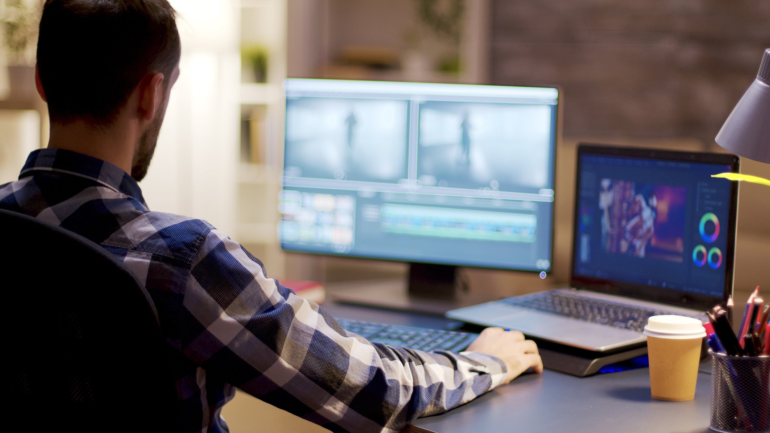 A man editing in video editing software on dual monitors in a home office with a laptop and a coffee cup on the desk.