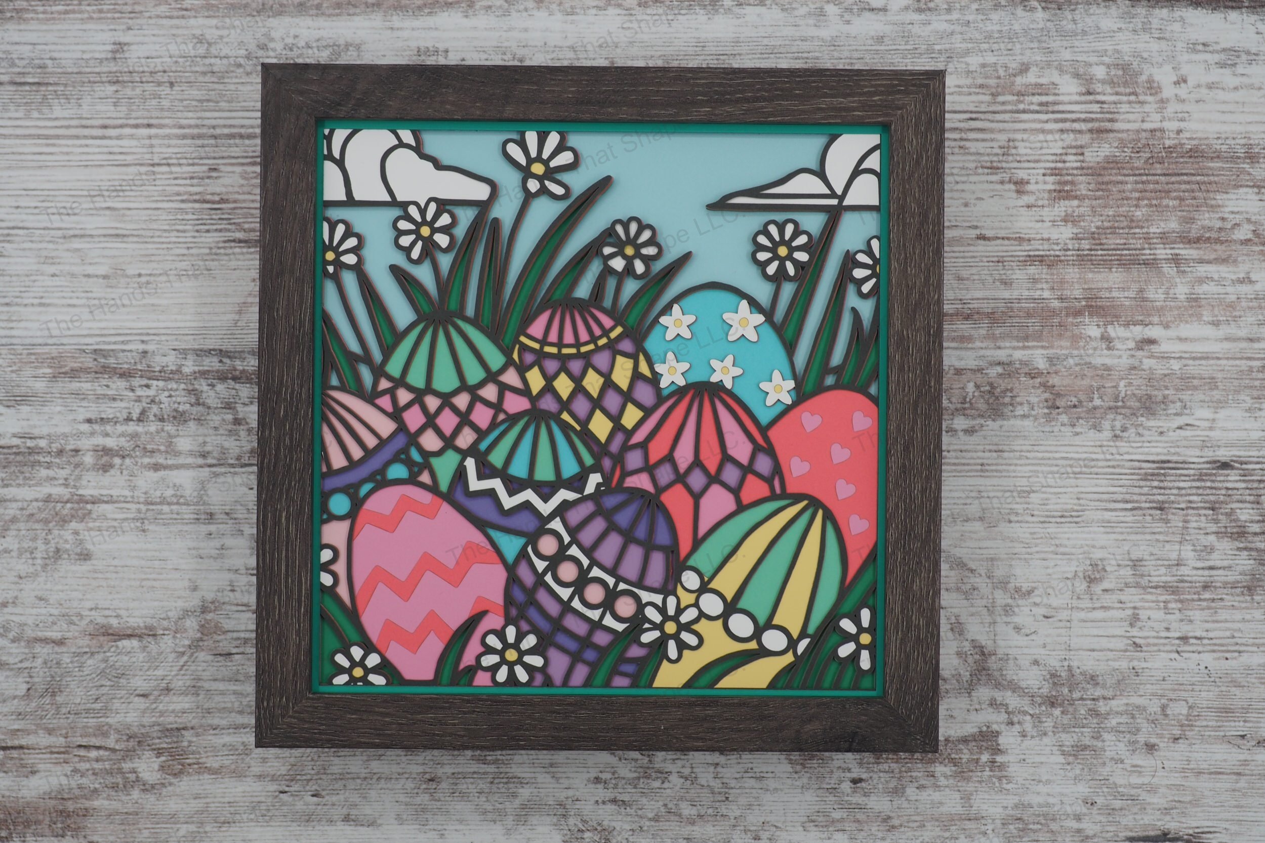 A vibrant layered paper shadowbox featuring intricately decorated Easter eggs nestled in lush grass, surrounded by blooming daisies and a cloud-filled sky.