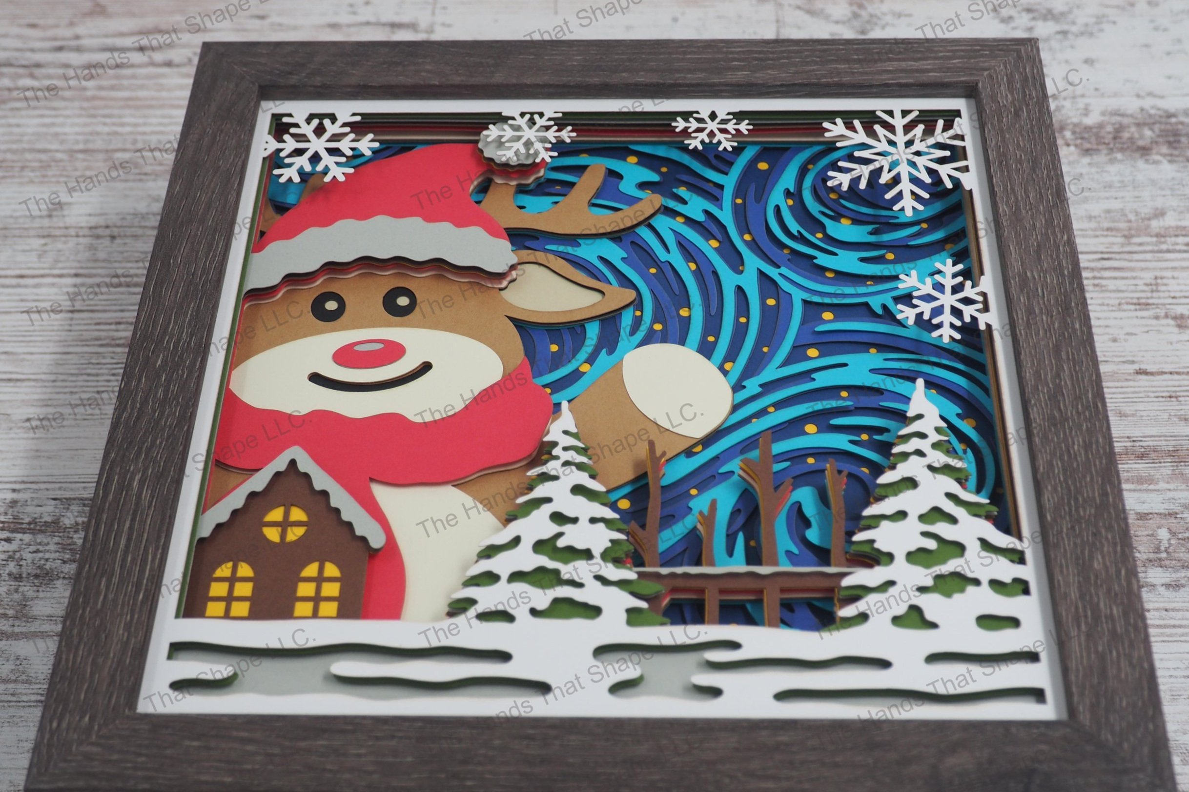A layered paper shadowbox depicting an oversized festive reindeer waving joyfully beside a snow-covered cabin, with wintery swirls and stars as a backdrop.