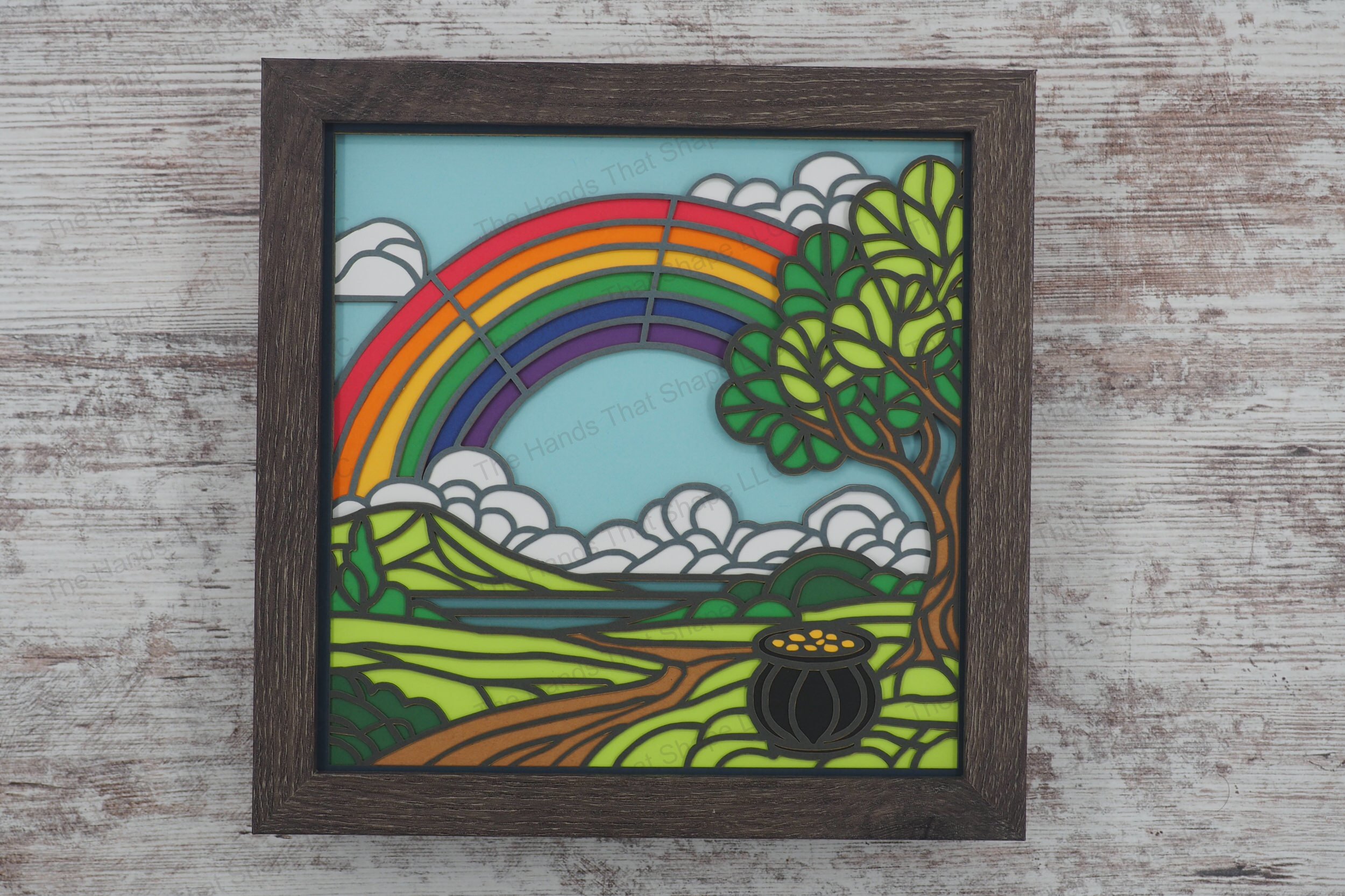 A vibrant layered paper shadowbox featuring a pot of gold, a winding path leading to a distant lake, and a brilliant rainbow arching across the sky.