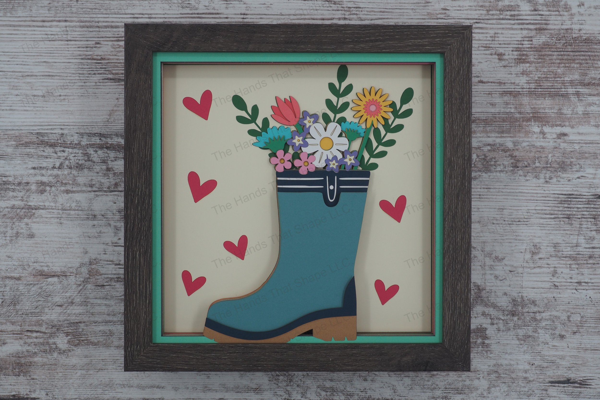 This springtime layered paper shadowbox features a bouquet of daisies, straw flowers, and lush greenery arranged playfully in a whimsical rain boot vase.