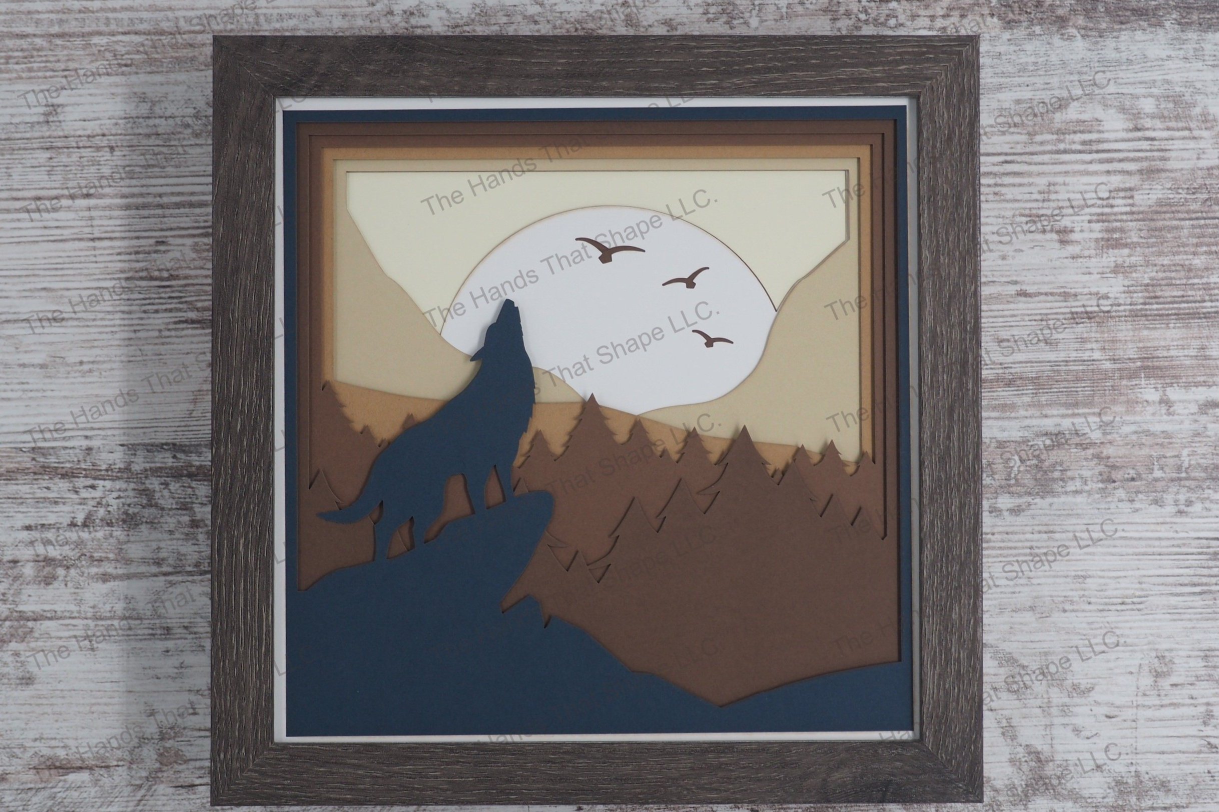 A framed layered paper shadowbox depicting a lone wolf howling at a full moon from a rocky ledge, with a backdrop of pine trees, rolling hills, and mountains.