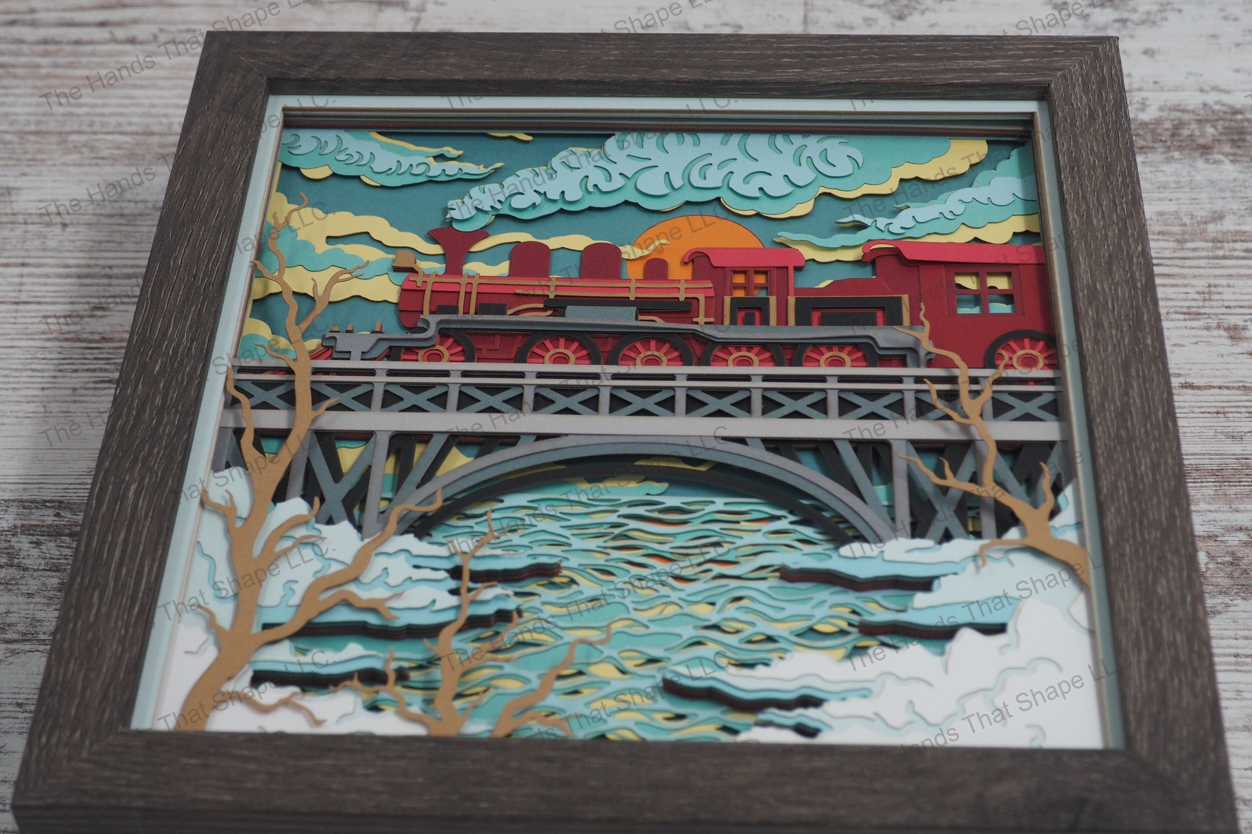 A detailed layered paper framed shadowbox depicting a classic steam train billowing smoke as it crosses a high bridge over a river, with a setting sun.