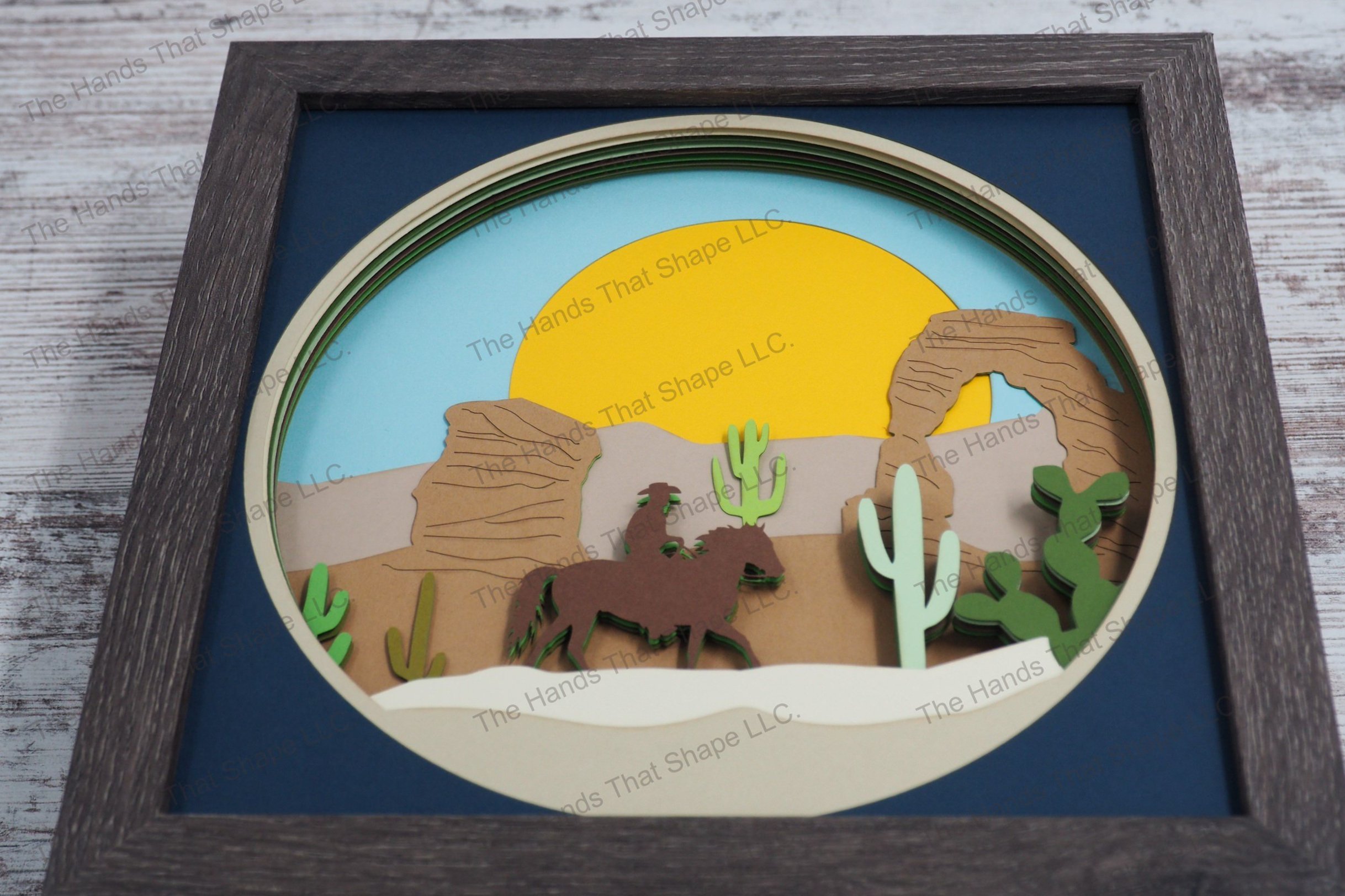 A striking depiction of a cowboy on horseback navigating through a desert landscape, this layered paper shadowbox features cacti, a stone arch, and sun.