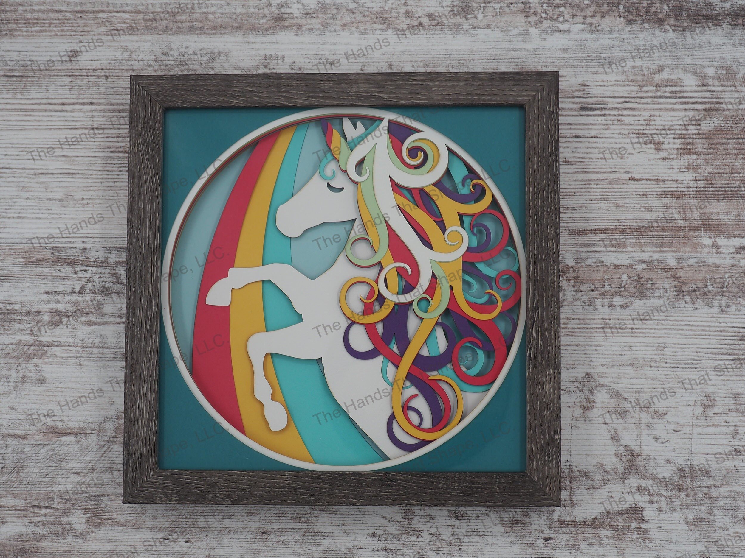 The layered paper shadow box features a white unicorn with a multi-colored mane prancing along a rainbow.