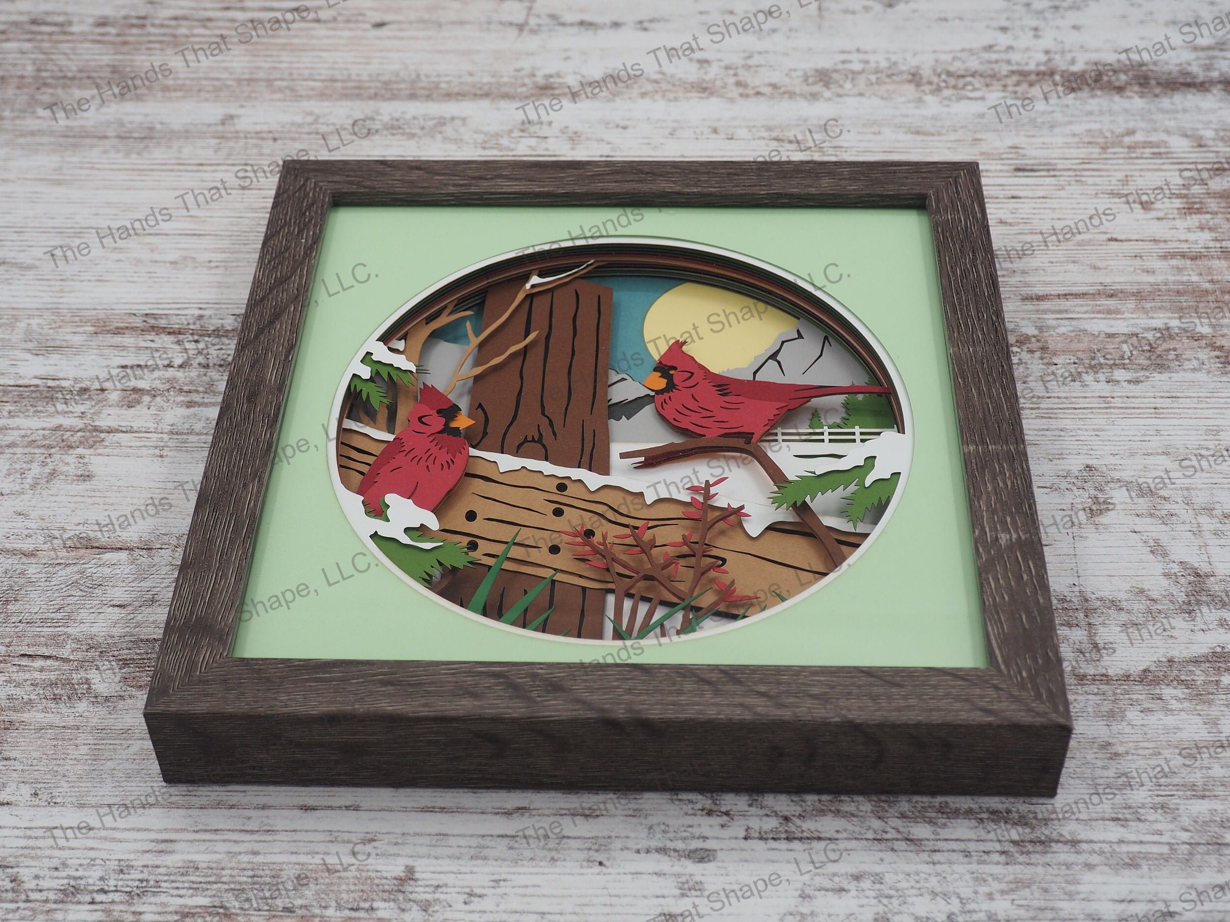 The layered paper shadow box features two colorful red cardinals perched upon a wooden fence and snowy background.