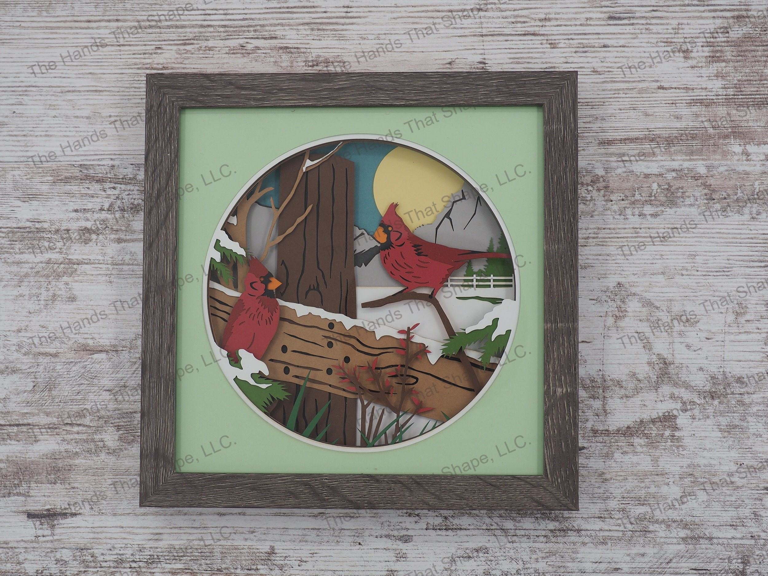 The layered paper shadow box features two colorful red cardinals perched upon a wooden fence and snowy background.