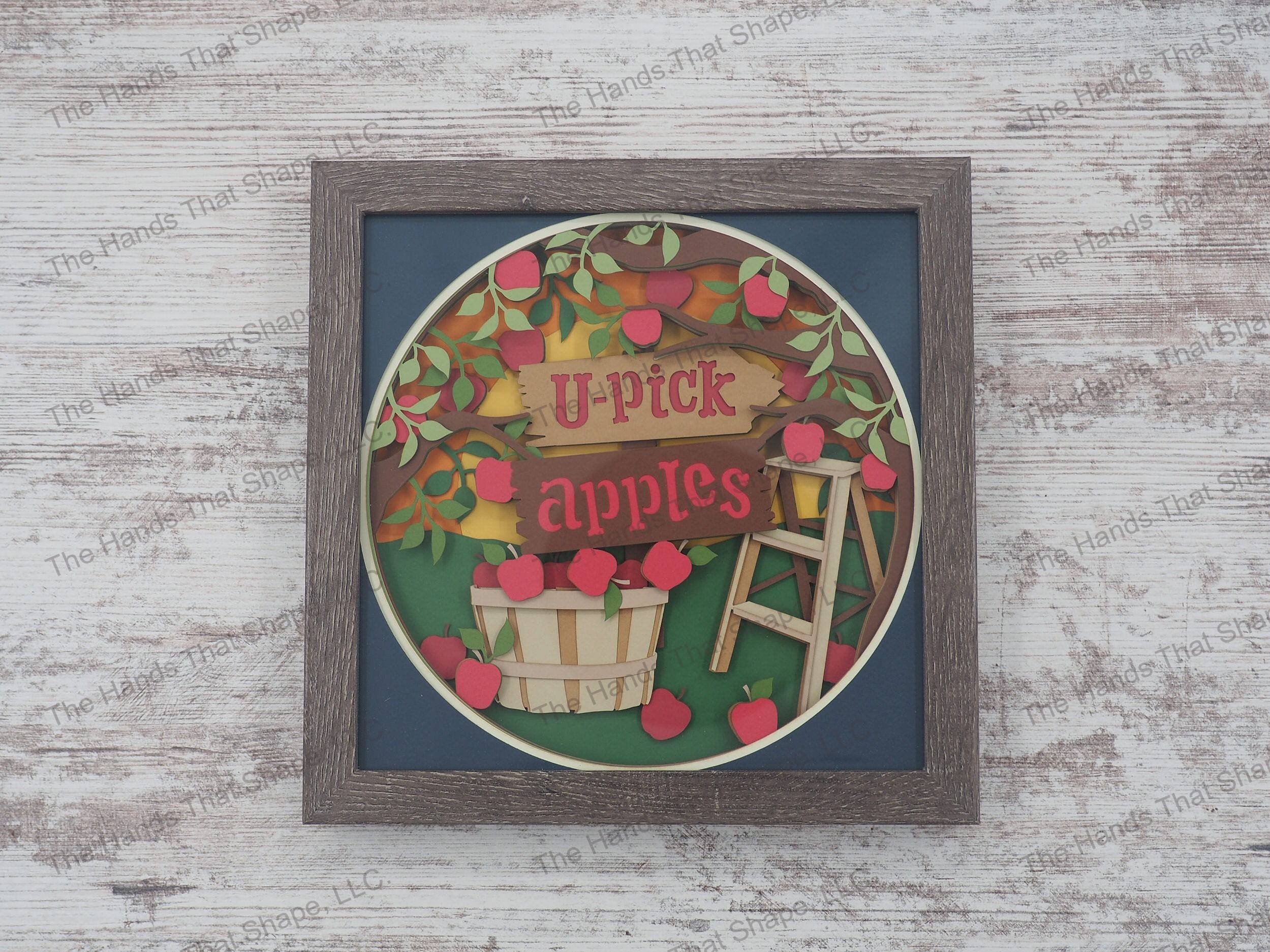 The shadow box features layers of cardstock paper to create a 3d bushel of fresh picked apples among an apple orchard.